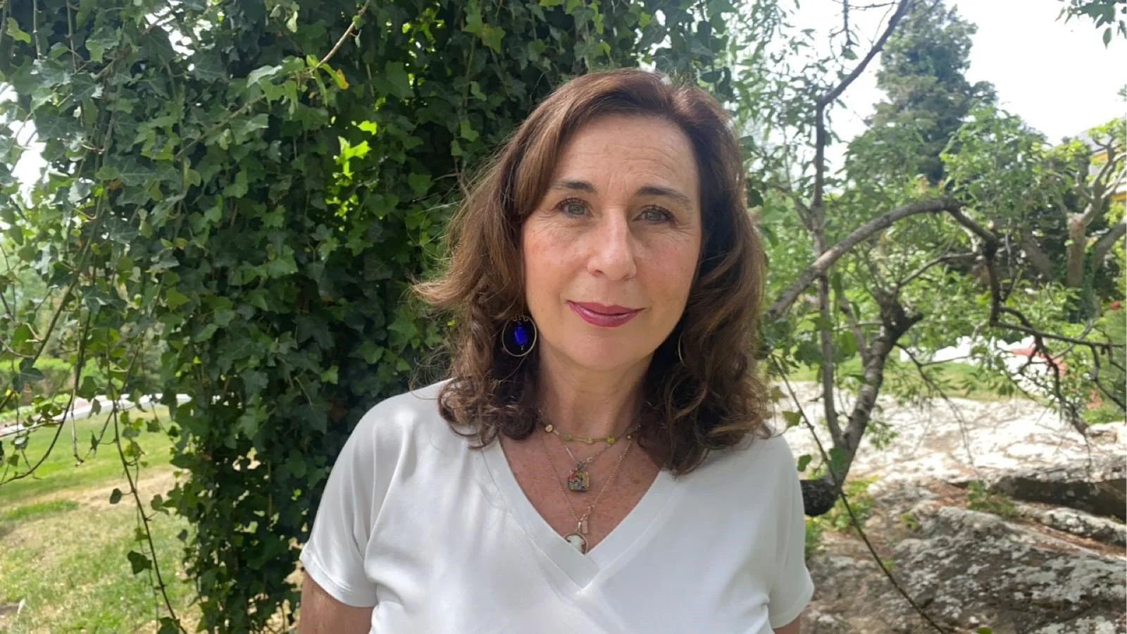 Mujer con cabello castaño y vestido blanco, con joyería colorida, de fondo natural con árboles y césped.
