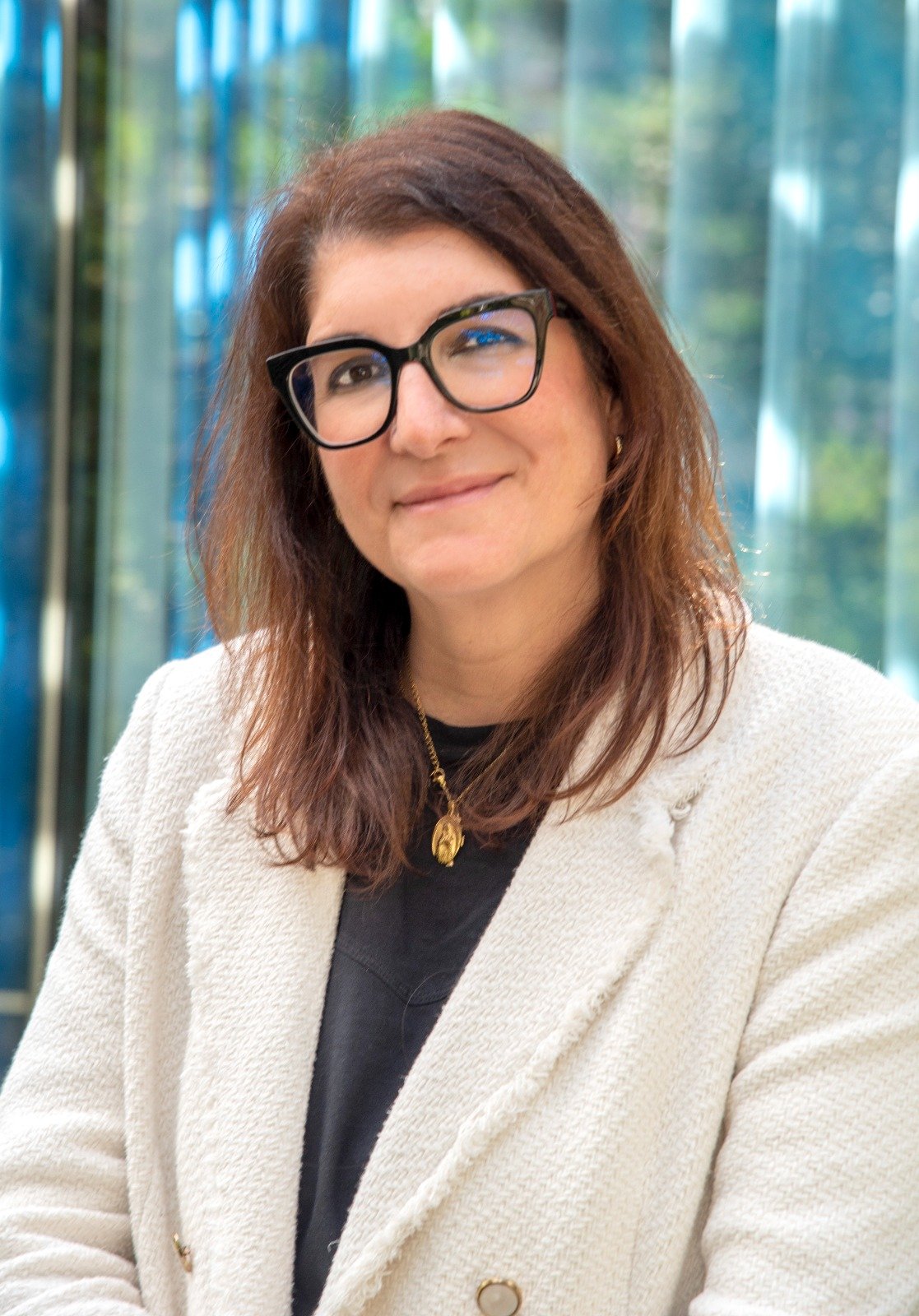 Mujer con cabello castaño y gafas negras posando en un entorno con fondo de cortinas translúcidas de color azul y verde.