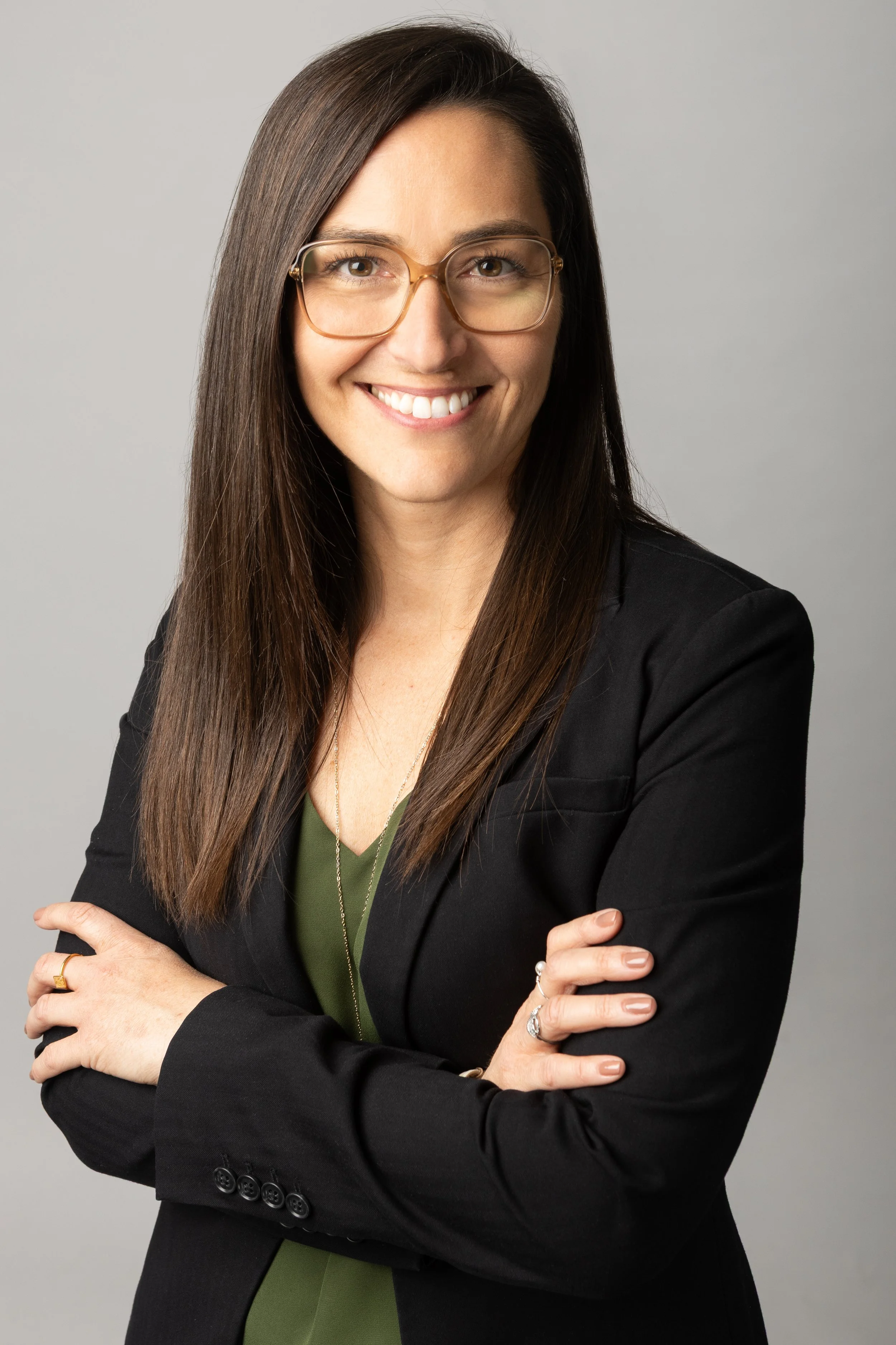Une femme souriante portant des lunettes élégantes, habillée d'un blazer noir et d'un haut vert, avec des cheveux longs et bruns, posant avec les bras croisés devant un fond gris.