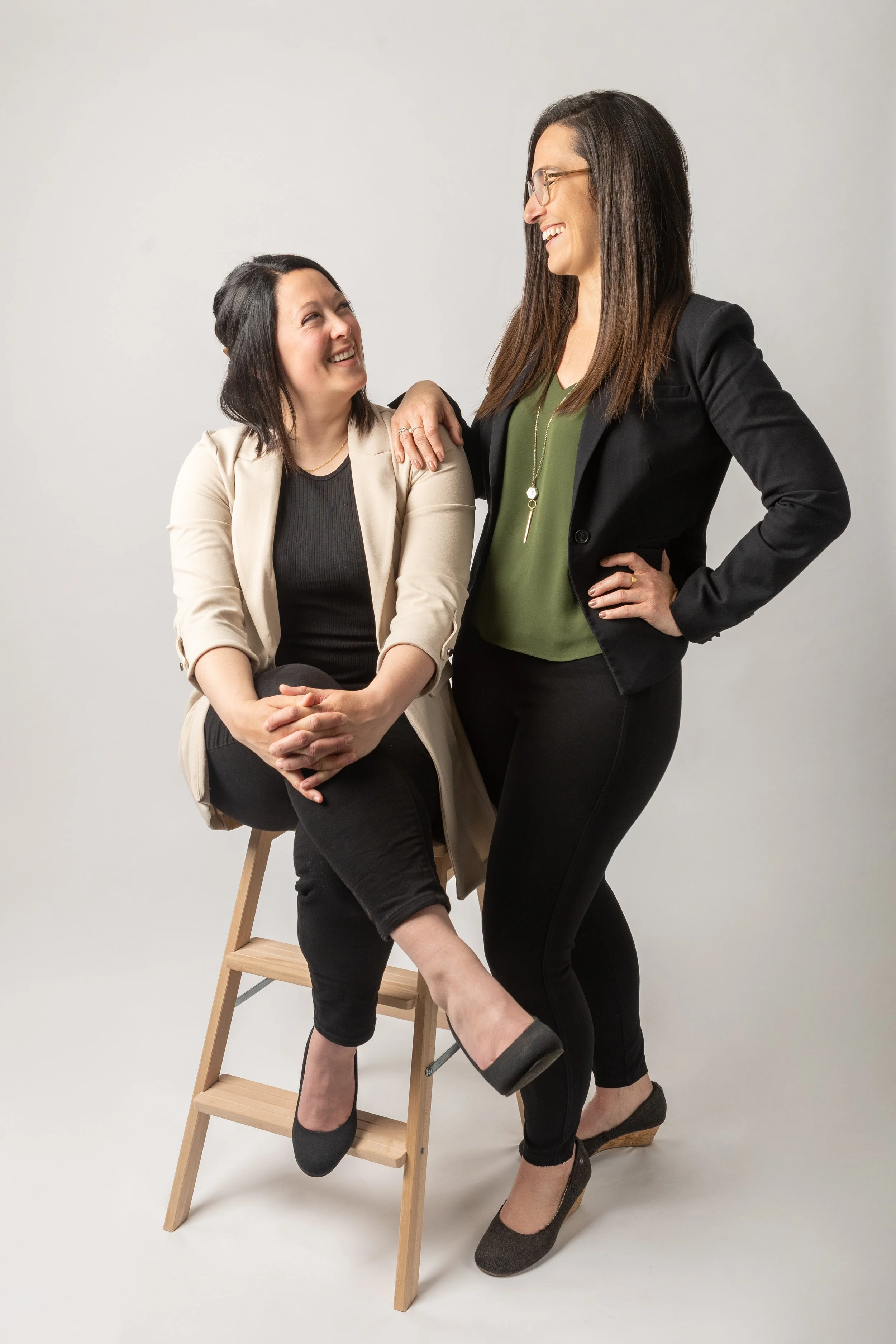Deux femmes souriantes échangent dans un studio avec un fond neutre. L'une est assise sur une chaise en bois, l'autre se tient à côté, posant sa main sur son épaule.