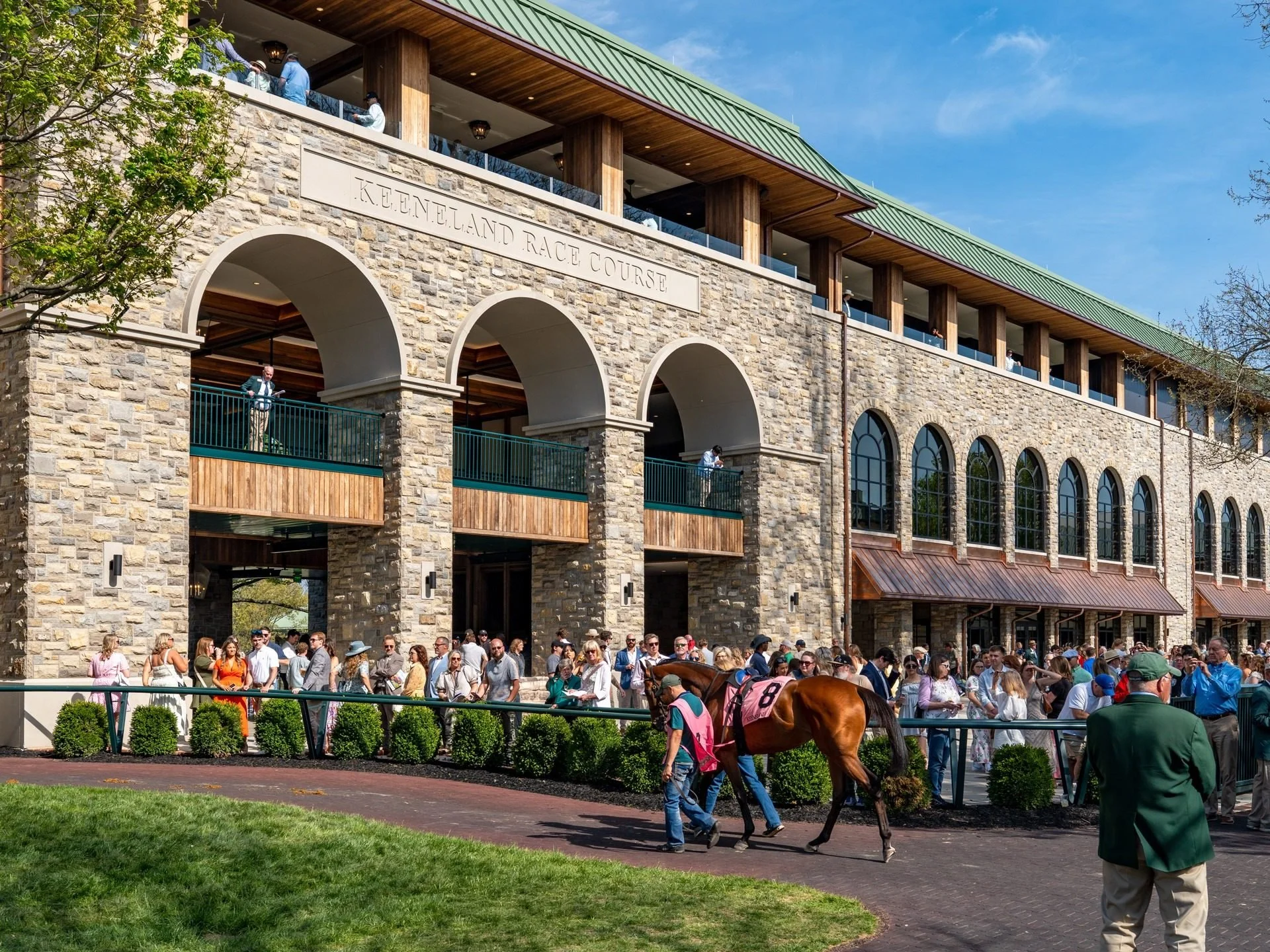 Crowd at Keeneland, with some people leading horses around the area, during a sunny day with a clear sky. The building features stone and wooden architecture with an arch and windows. The scene appears lively with attendees at a horse racing event.