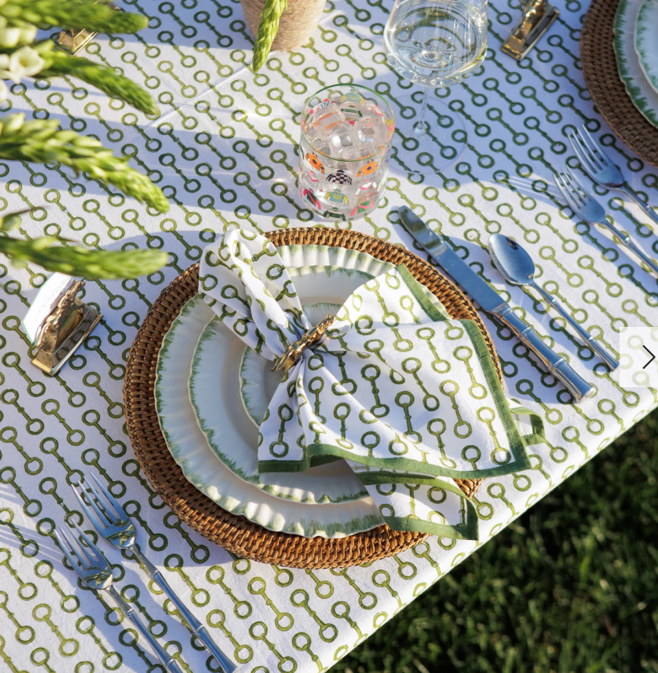 A neatly set outdoor dining table with green and white key-patterned tablecloth, plates with green and white rimmed design, napkins folded on plates, glass of water, and silverware.