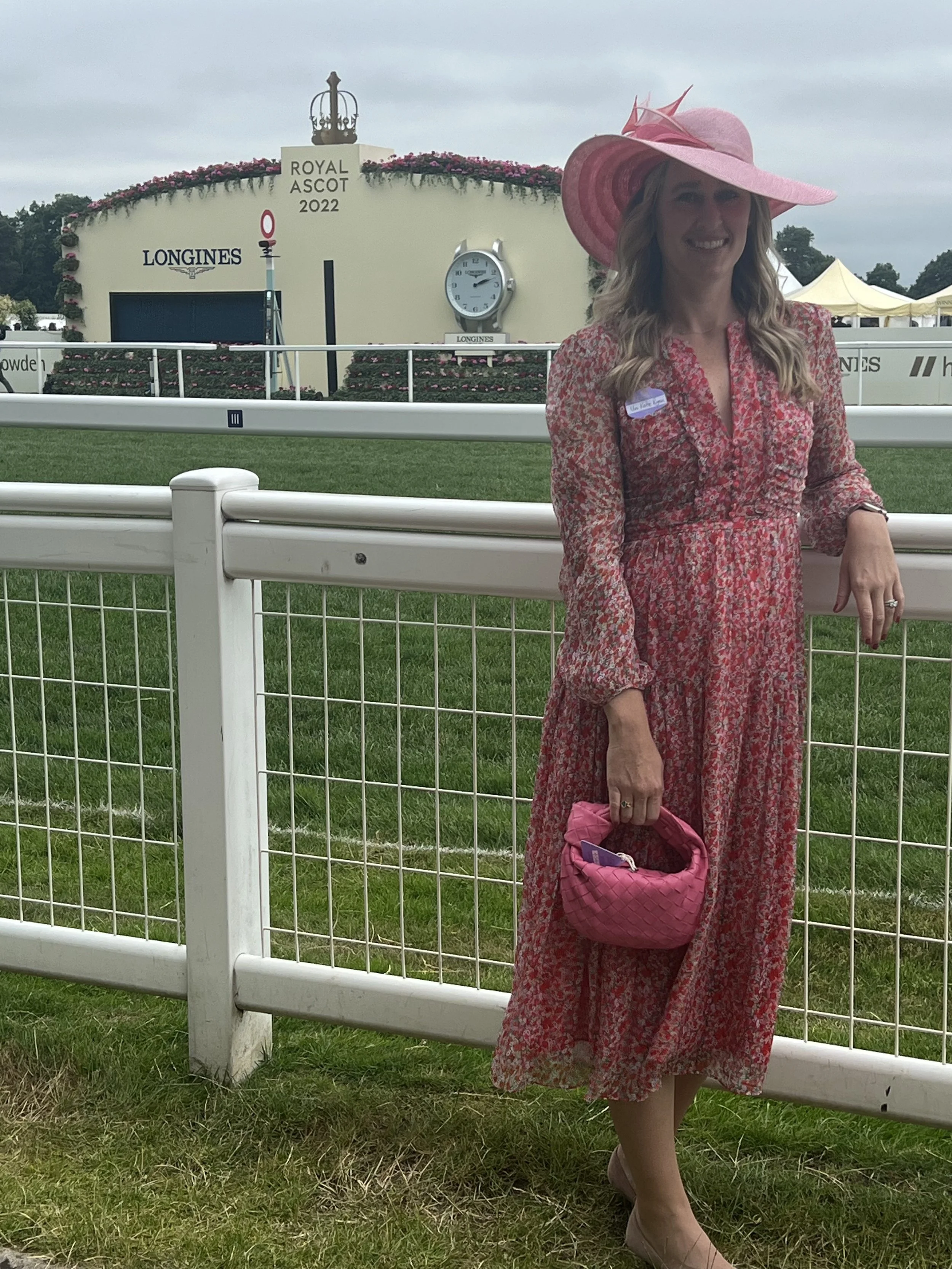 A woman in a pink floral dress and wide-brimmed pink hat smiling at the camera, holding a pink quilted handbag, standing in front of a white fence at a horse racing event with the sign 'Royal Ascot 2022' in the background.