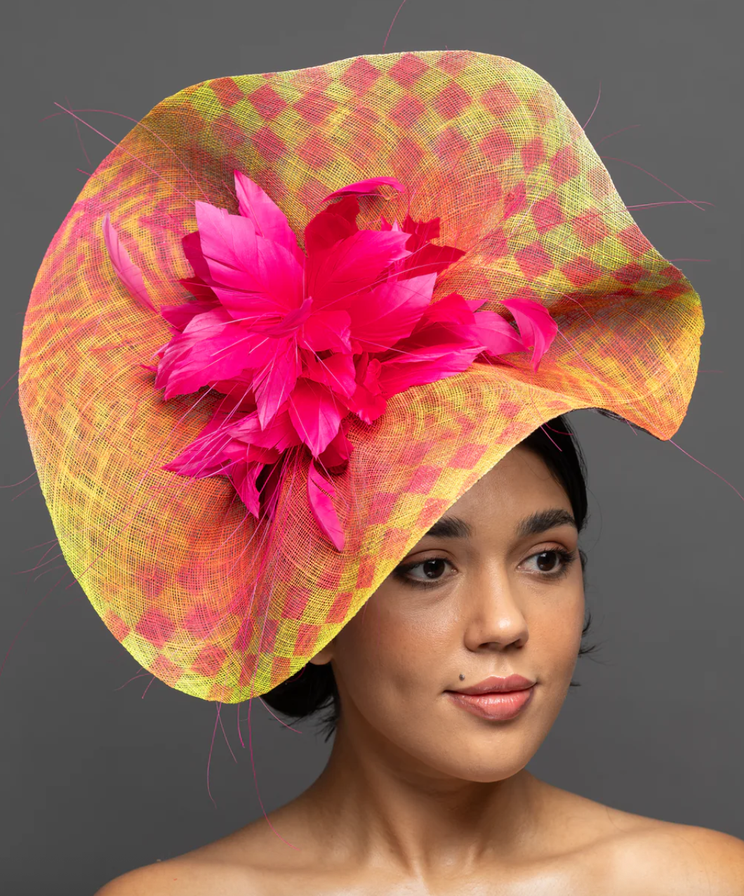 A woman with short dark hair wearing a large, colorful straw hat with wide brim. The hat is yellow with pink checks and decorated with large pink feathery flowers. Gray background.