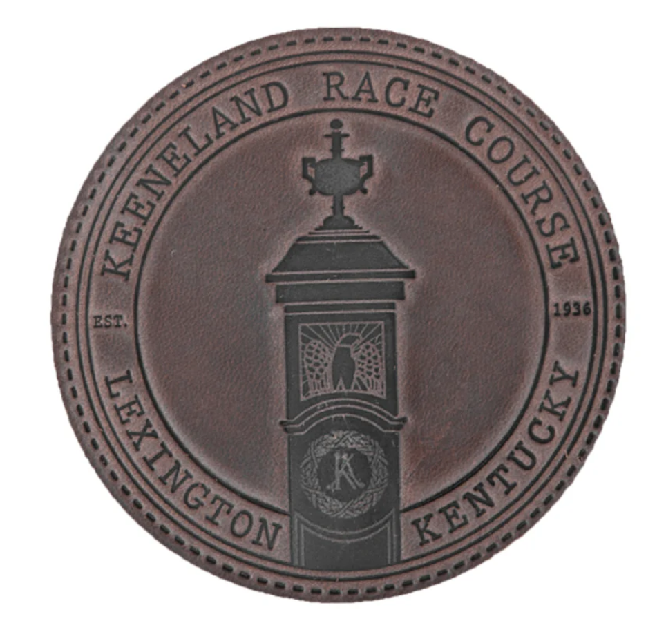 Close-up of a round coin or medallion with the inscription 'Kentucky Enetland Race Course Lexington Kentucky' and the year 1936. The center features a clock tower with a cross on top, a butterfly flower stained glass window, and a circular emblem with the letter 'K'.