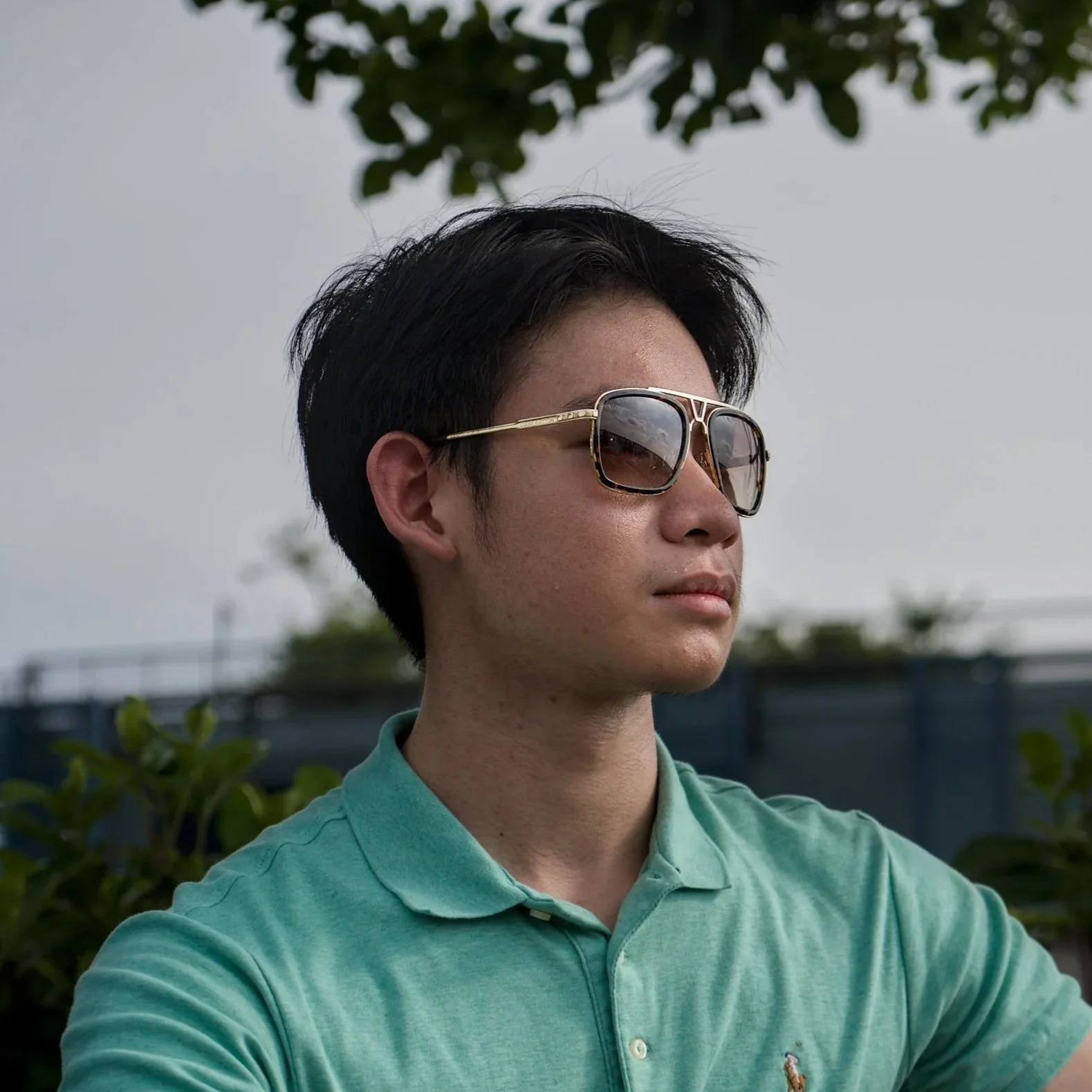 A young man wearing sunglasses and a mint green polo shirt outdoors with greenery and a fence in the background.
