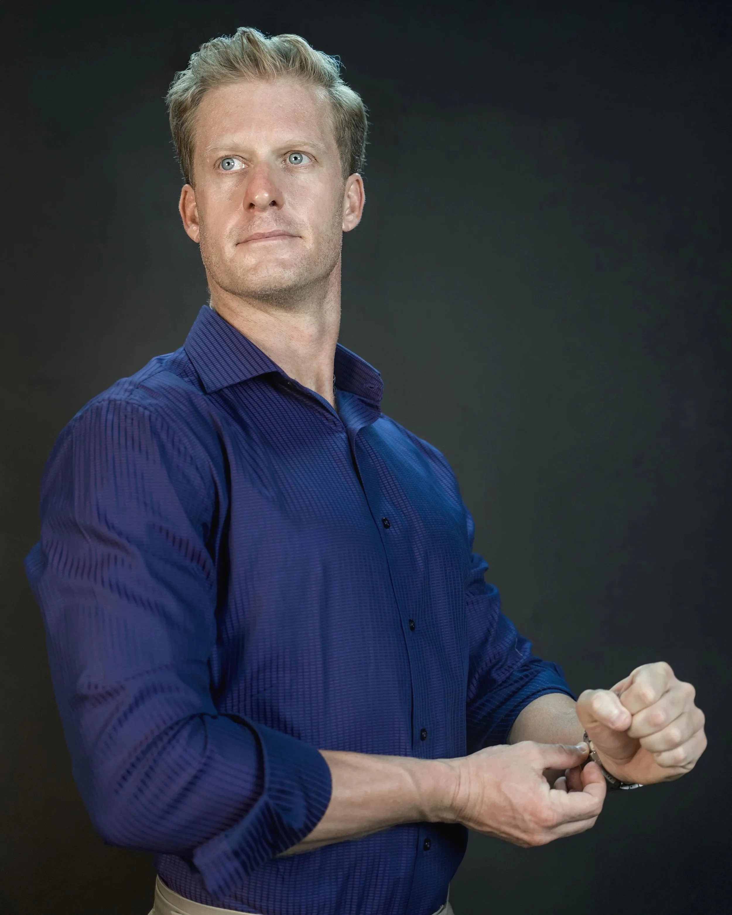 A man with blonde hair and blue eyes wearing a dark blue checked shirt, standing against a dark background, with his arms crossed and hands clasped.