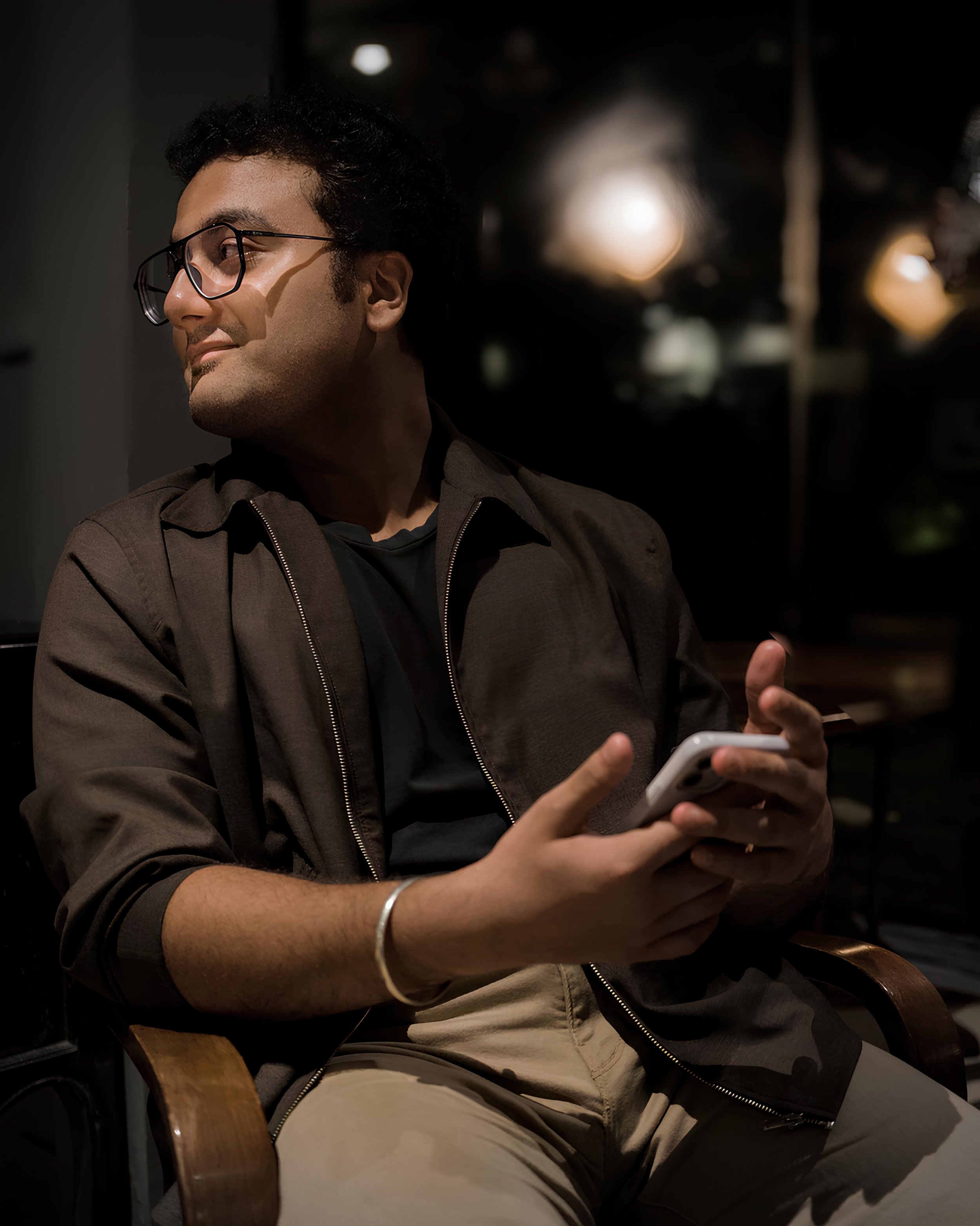 A young man with glasses sitting in a dimly lit area, looking to the side while holding a smartphone in his hands.