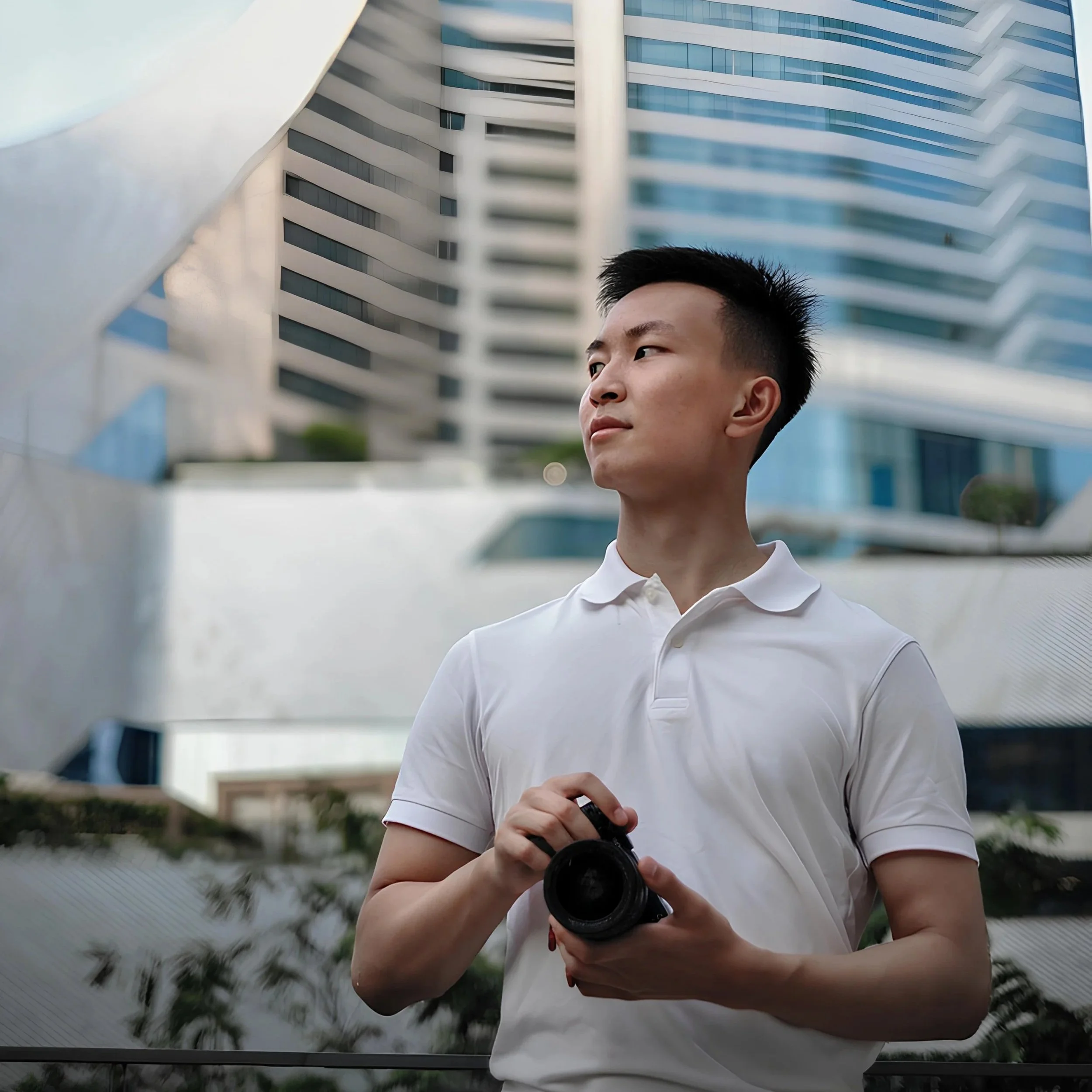 A young man with short black hair wearing a white polo shirt, holding a camera in his hands, standing outdoors in front of modern high-rise buildings, with some greenery and a water feature nearby.