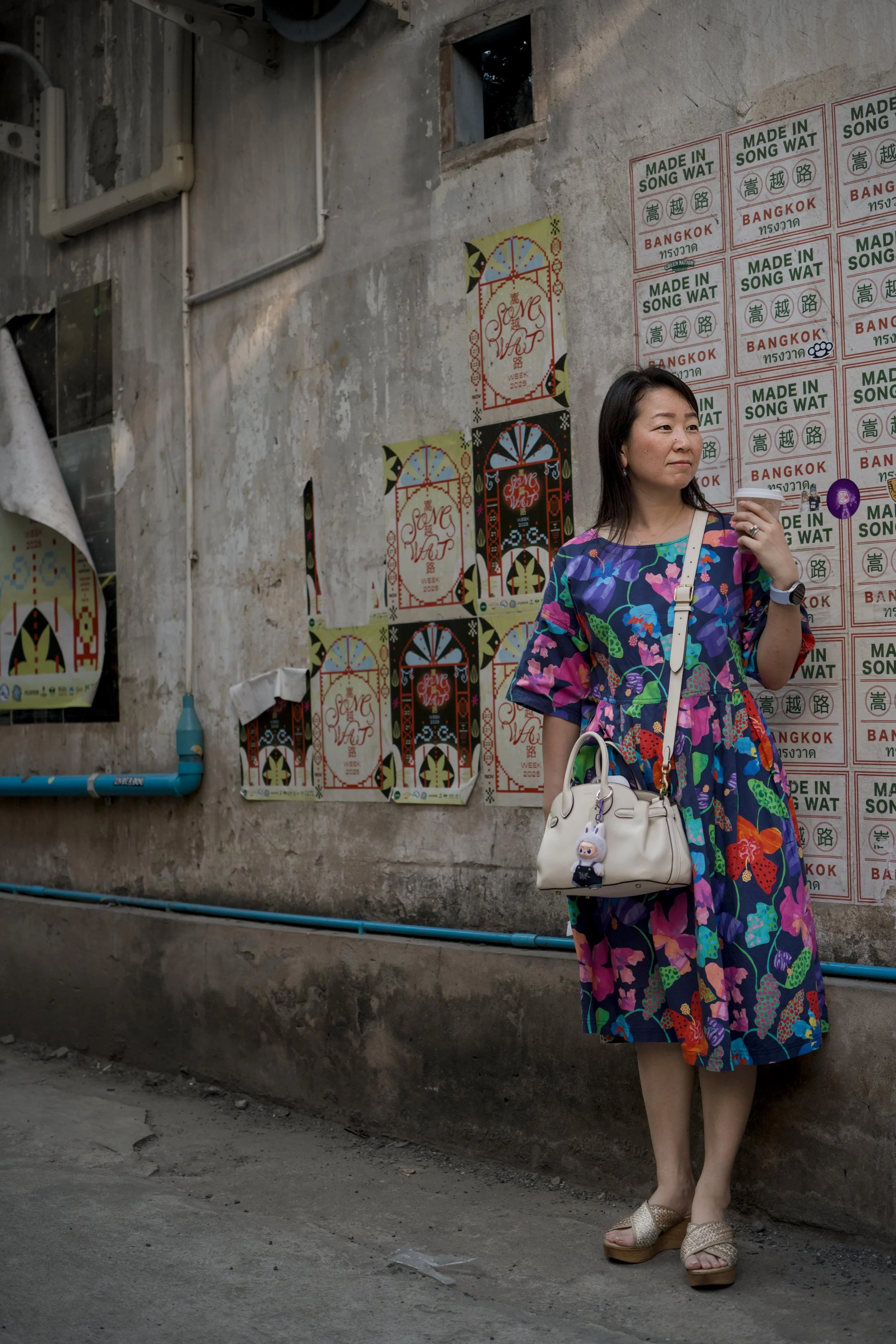 A woman in a colorful dress holding a white purse and a drink, standing against a weathered wall with posters that read 'Made in Song Wat' and 'Bangkok', with painted pipes below.