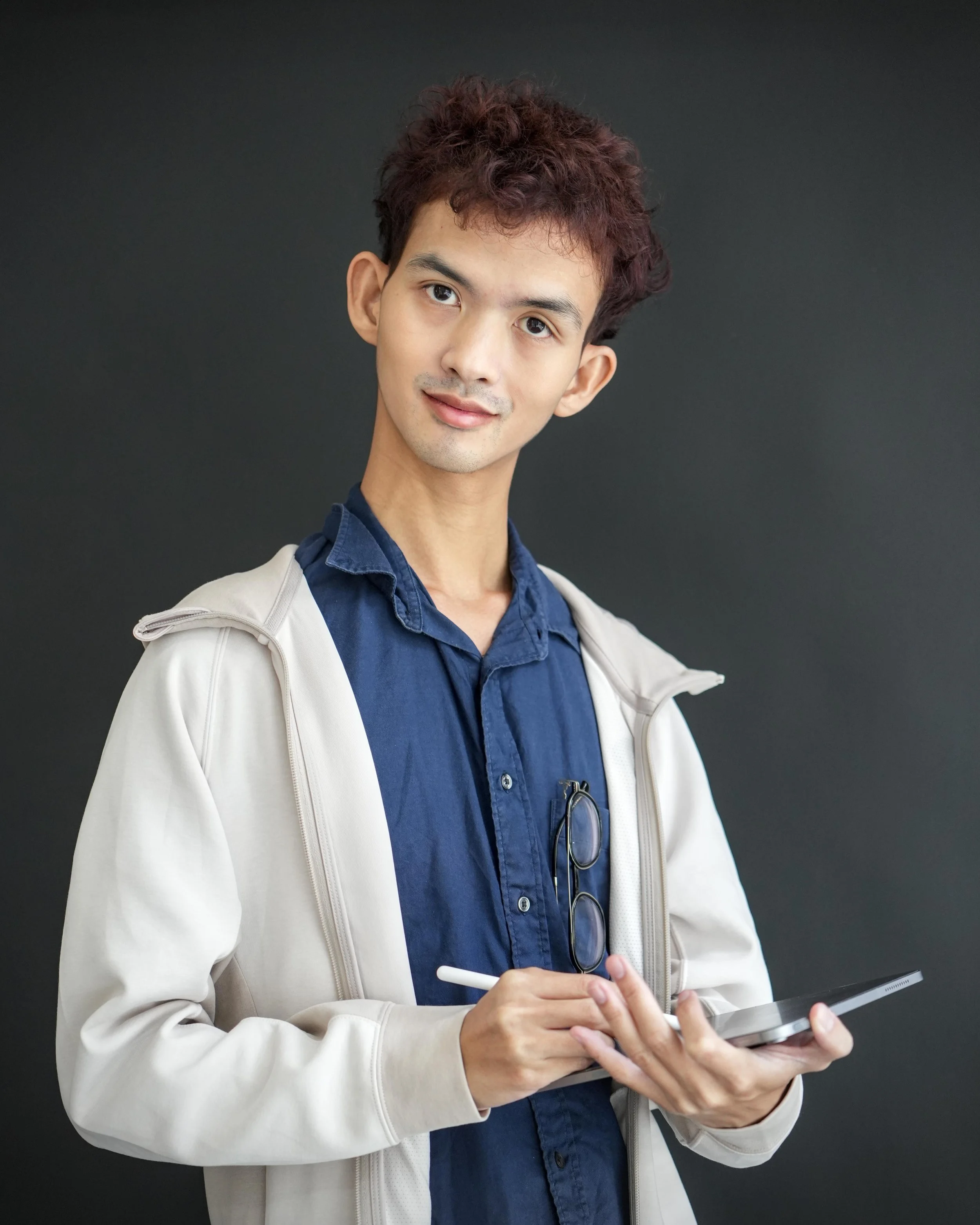 A young man with curly brown hair and light skin, dressed in a blue shirt and beige jacket, holding a tablet and looking into the camera with a slight smile.