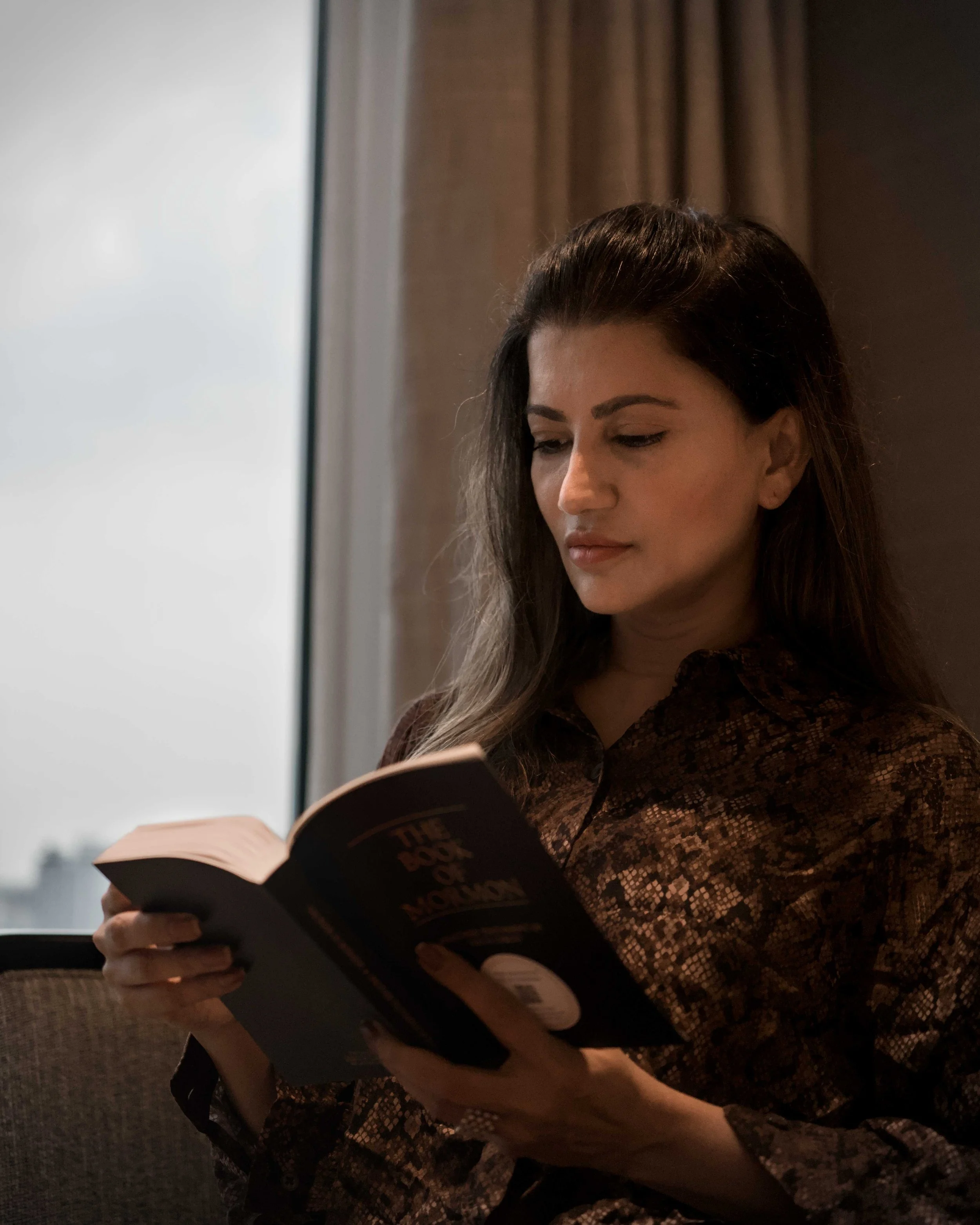 A woman with dark brown hair and light skin reading a book while sitting in front of a window with brown curtains.