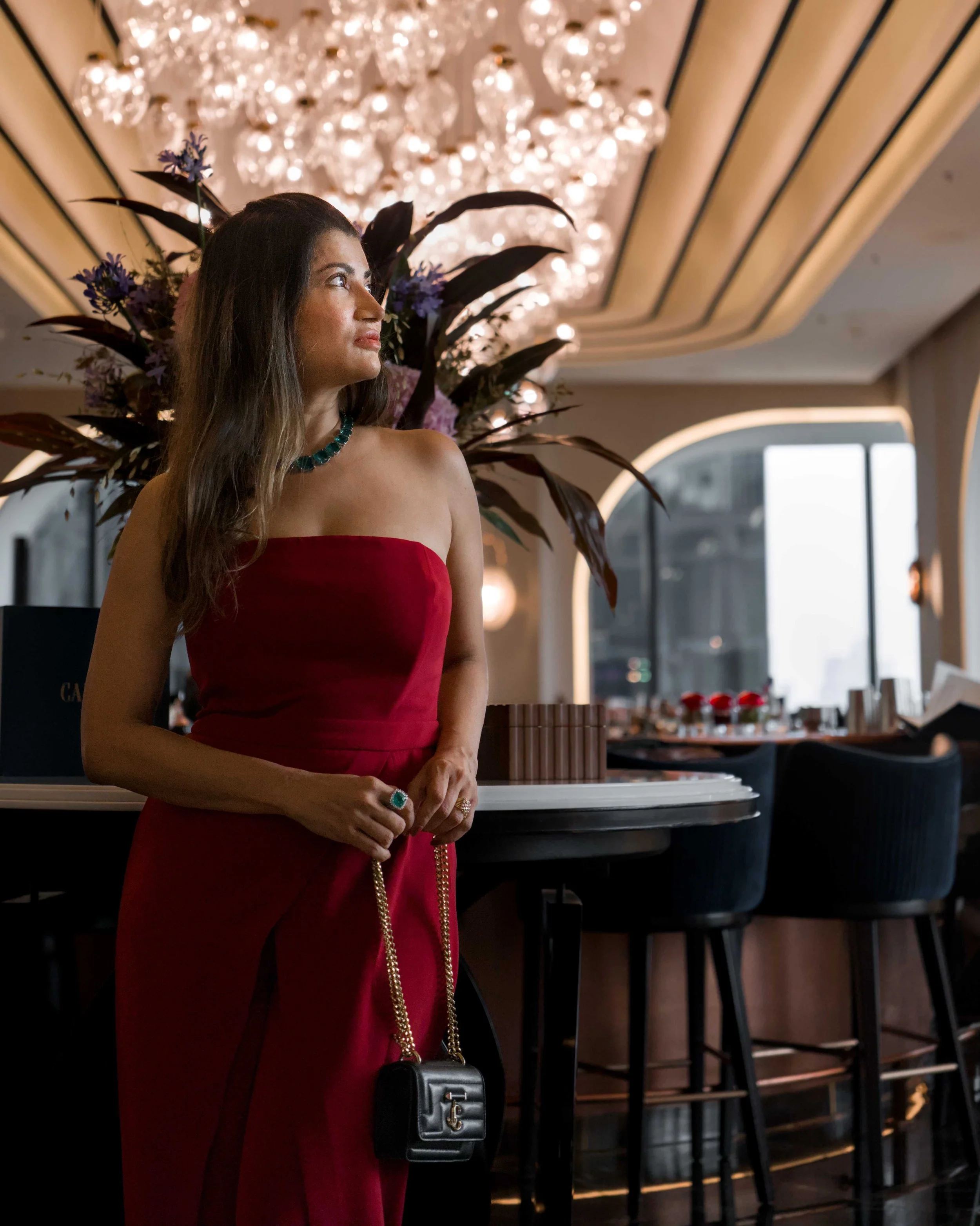 A woman in a strapless red dress standing inside a room, with ornate ceiling design and a large chandelier hanging above her. She has long dark hair, wears a green necklace, a ring, and holds a small black handbag with a gold chain. There are high ta