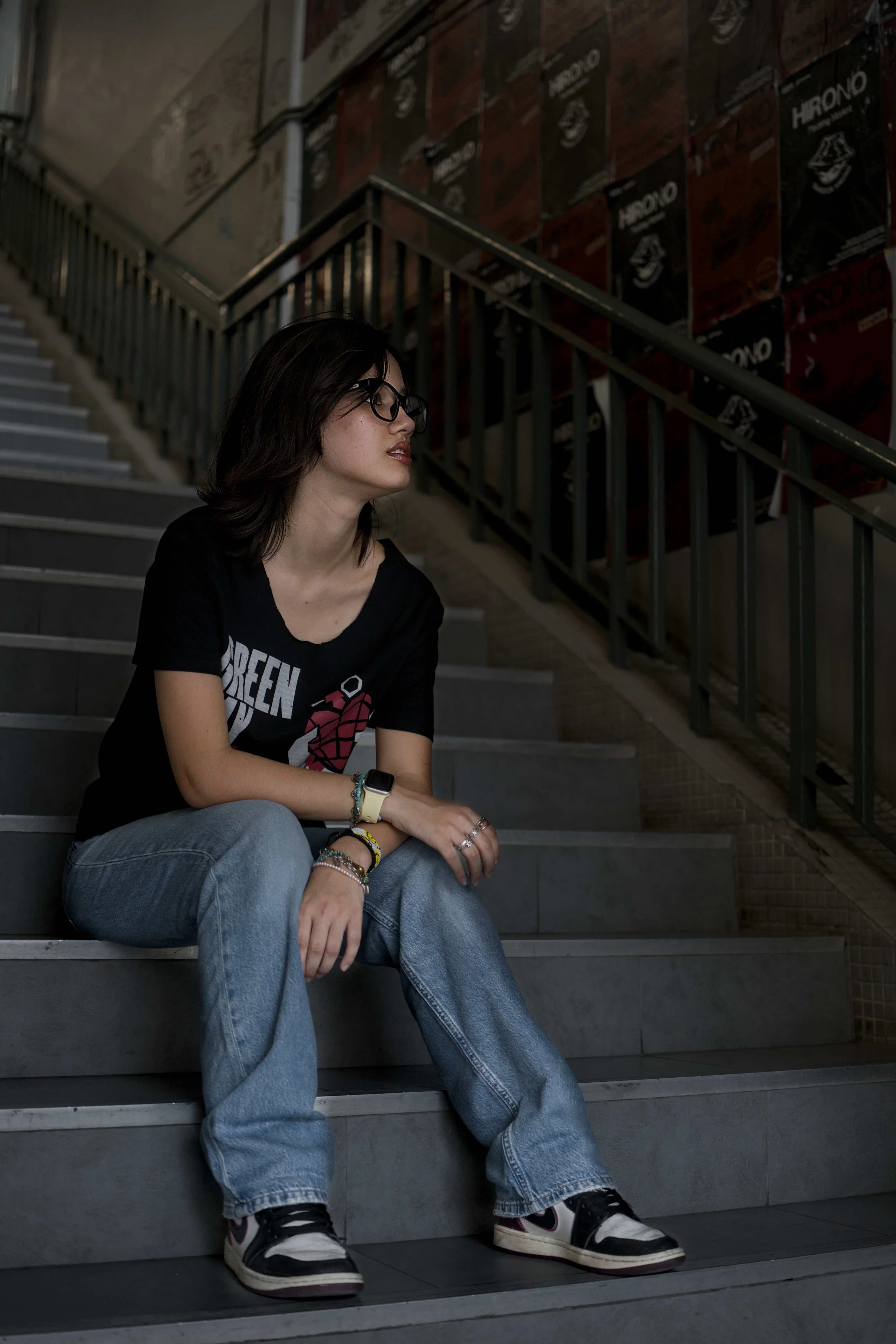 A young woman with dark hair, glasses, wearing a black t-shirt with a graphic, light blue jeans, and sneakers, sitting on indoor stairs near a black railing and a wall covered with posters.