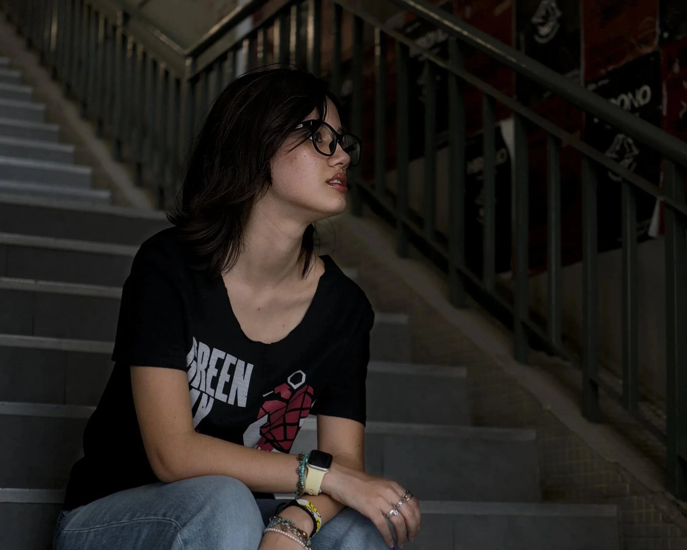 A young woman with dark, shoulder-length hair and glasses sits on a staircase, looking to her right. She is wearing a black t-shirt with red and white text and image, and has several colorful bracelets and rings on her left arm and hand. The background features industrial-style metal stairs and railings, with signs visible on the upper part of the wall.