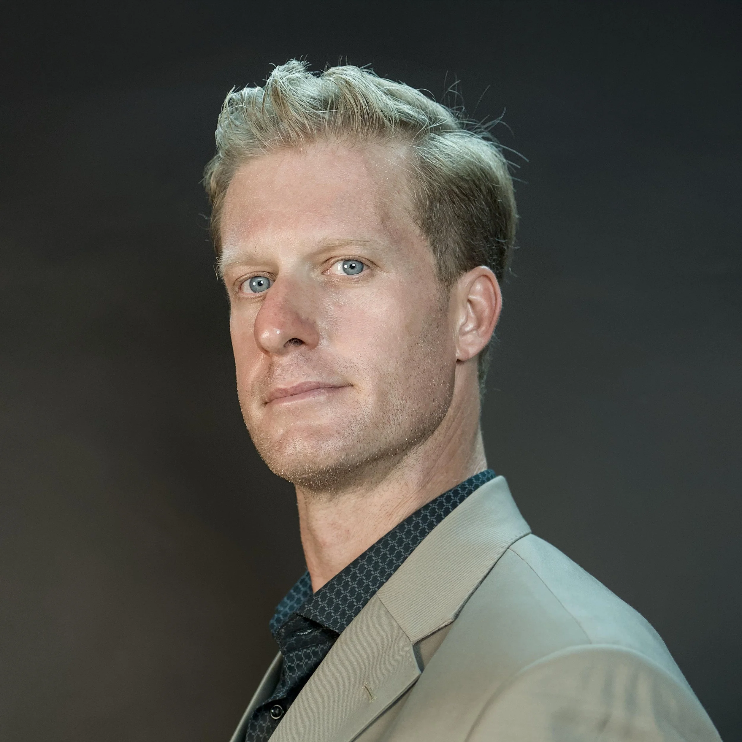 Close-up of a man with blue eyes, blonde hair, dressed in a beige suit jacket with a patterned shirt underneath, against a dark background.