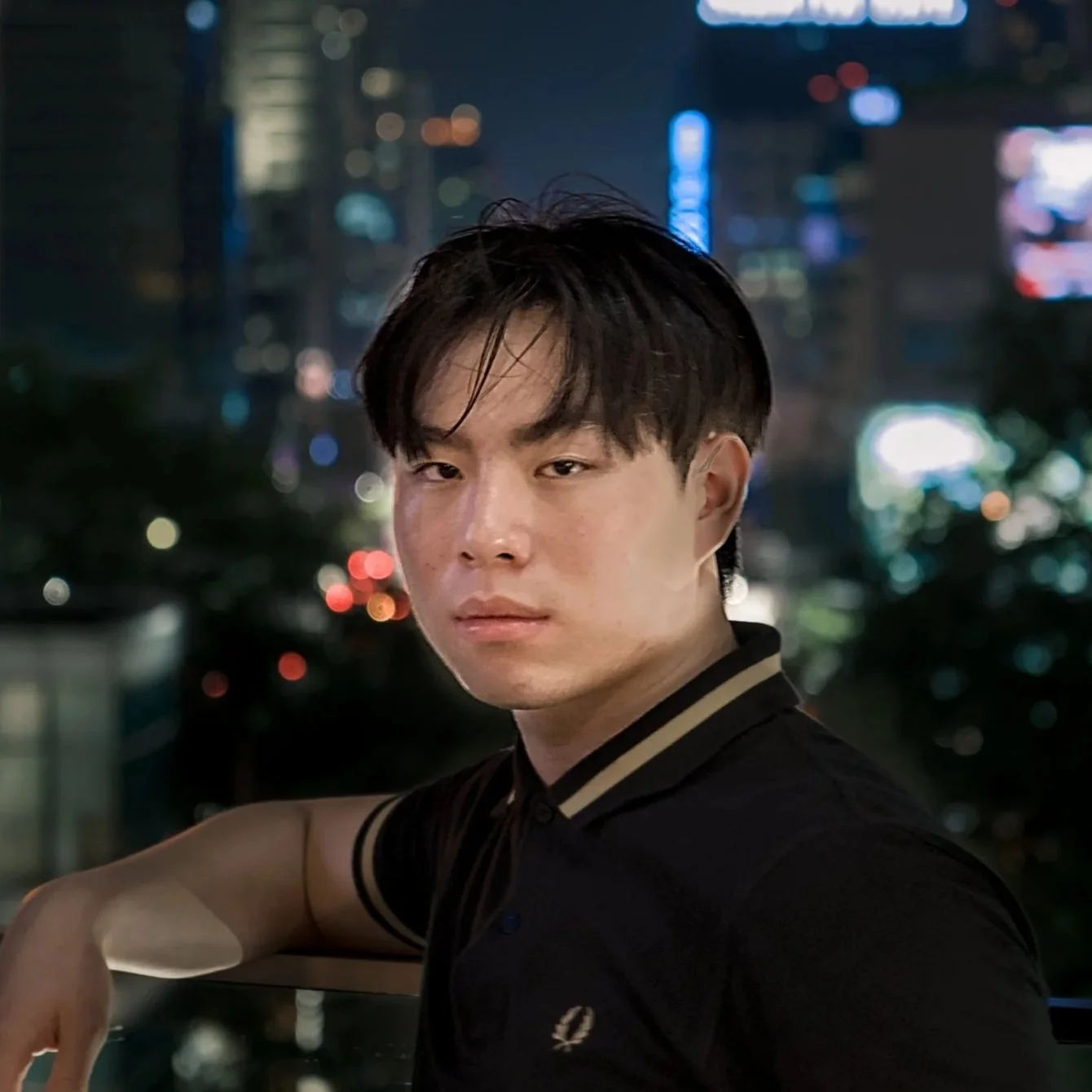 A young man with dark hair and light skin posing at night with a cityscape background of illuminated buildings and colorful lights.