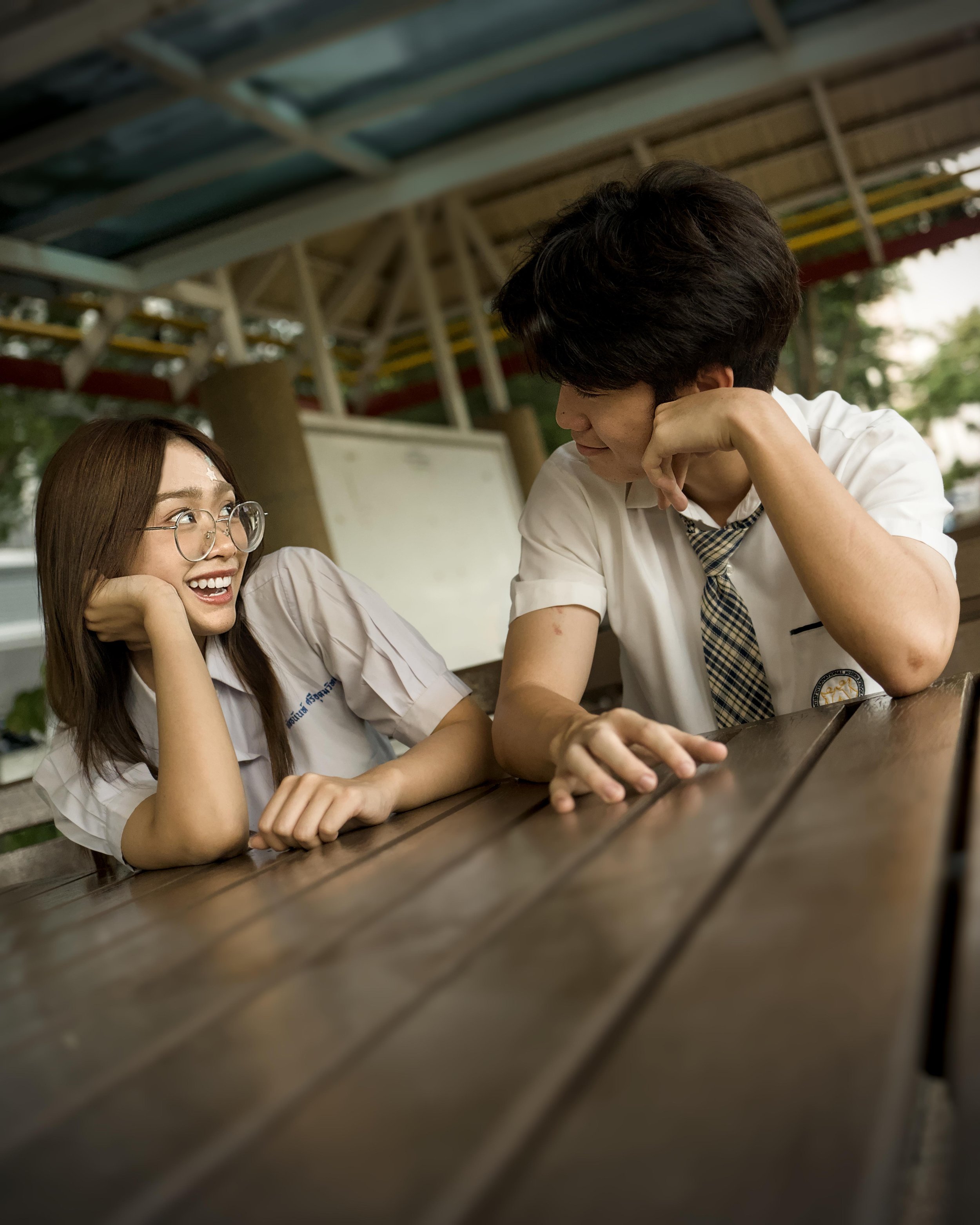 Smiling female student with glasses and long hair leaning on her hand, engaging with a male student with dark hair and a school uniform, at a wooden table outdoors under a colorful roof structure.