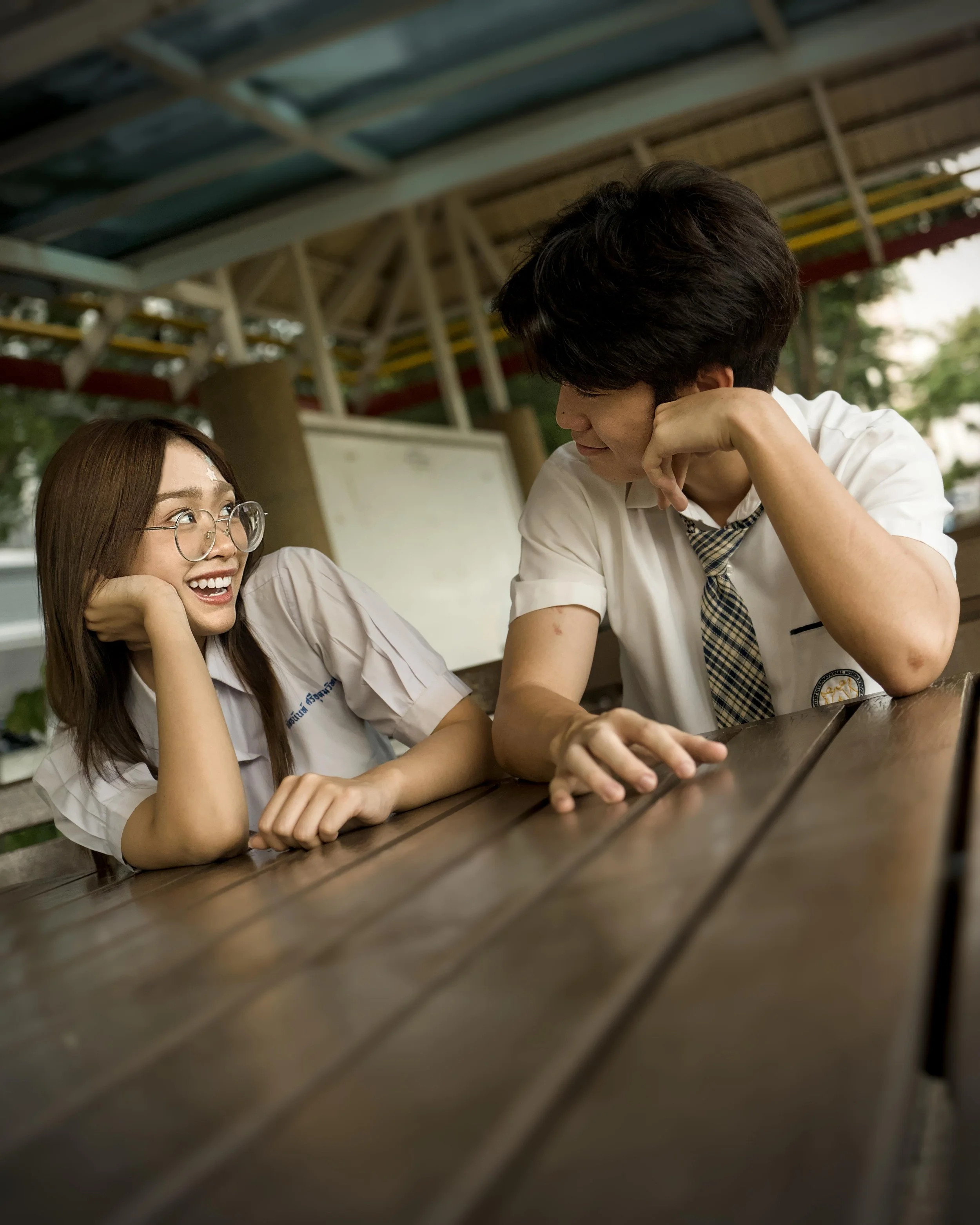 A woman with glasses and long hair smiling with her hand under her chin, looking at a young man with dark hair who is resting his chin on his hand and looking at her, both seated at a wooden table outdoors under a covered structure.