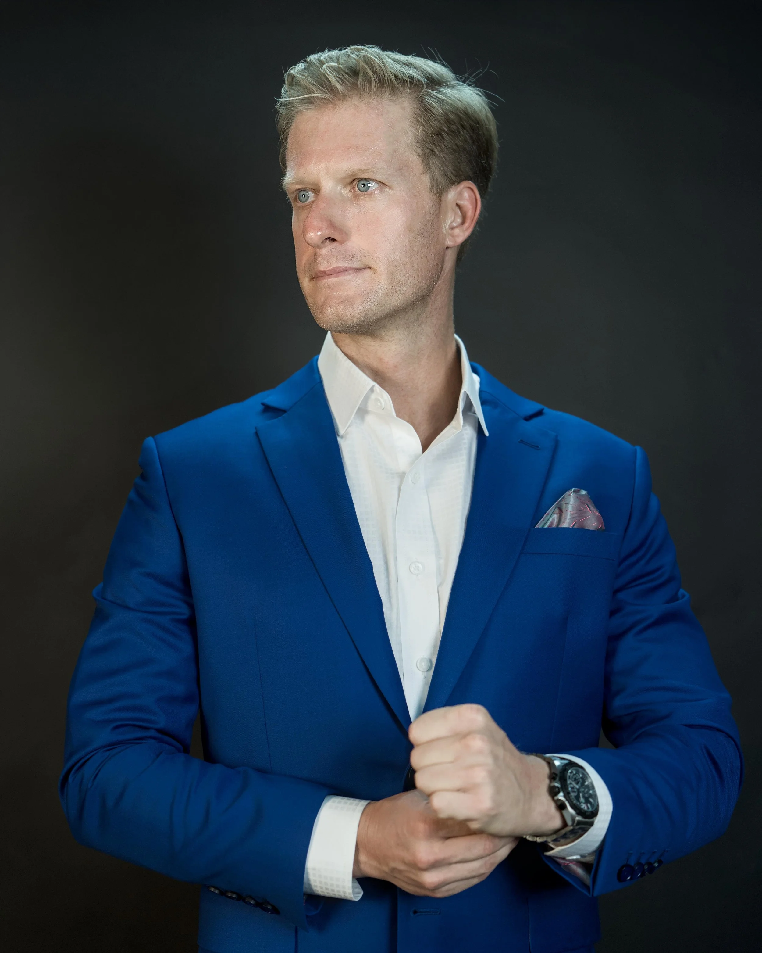 A man wearing a blue suit with a white shirt and a patterned pocket square, posing with his arms crossed against a dark background.