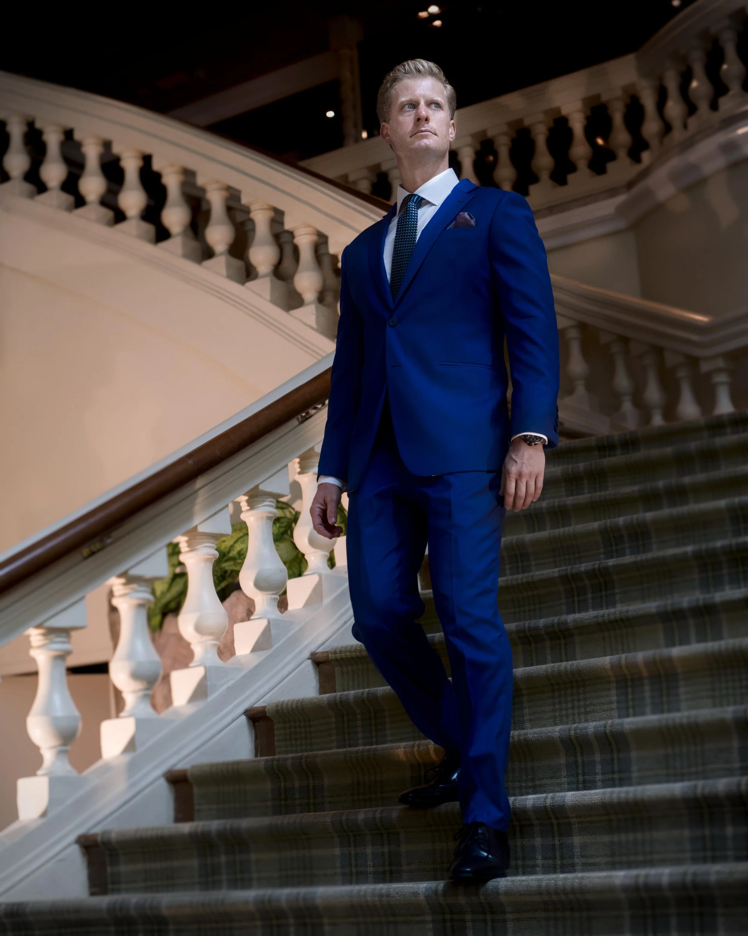A man in a blue suit and tie walking down a staircase with a decorative white balustrade in an elegant building.