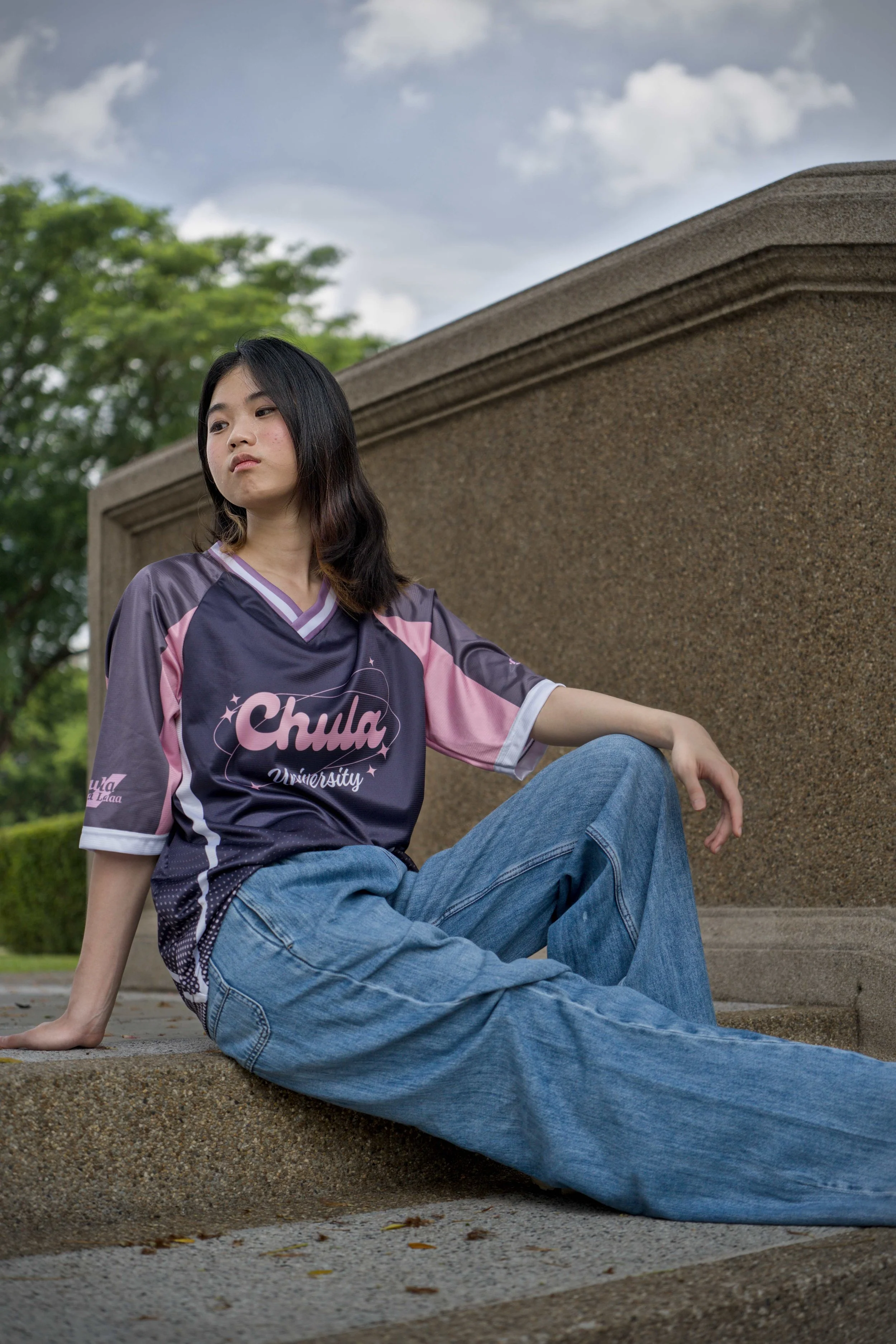 Young woman sitting on concrete steps outdoors, wearing a purple and pink sports jersey with the words "Chula University" on it, blue jeans, and has long black hair. She looks to the side with a serious expression, amid a background of trees and a cl