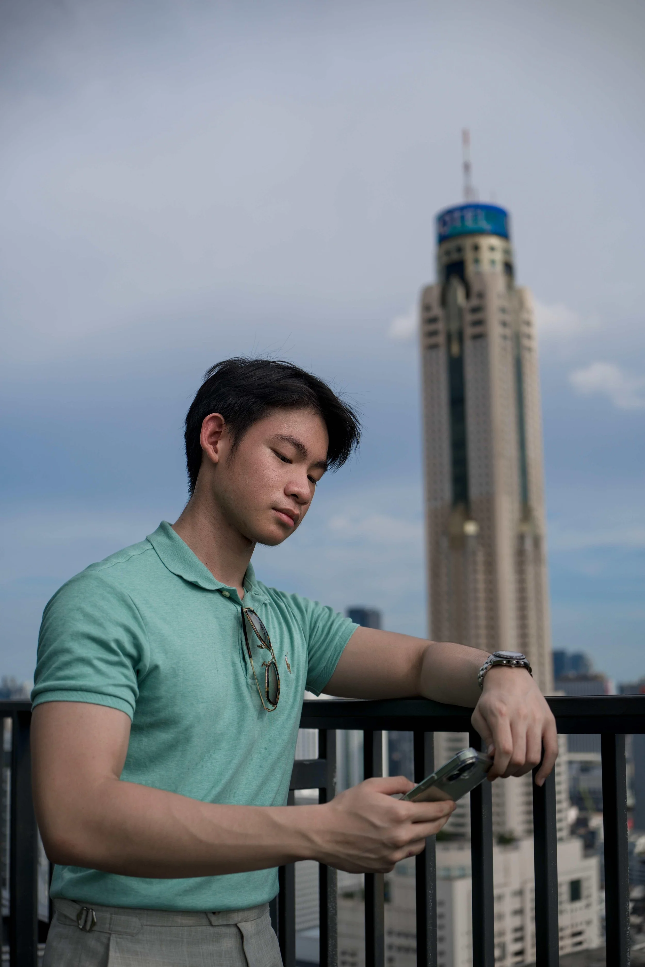 A young man with dark hair wearing a light green polo shirt and glasses hanging from his collar is looking at his phone on a balcony. In the background, there is a cityscape with tall buildings, including a skyscraper with a blue top, under a partly 