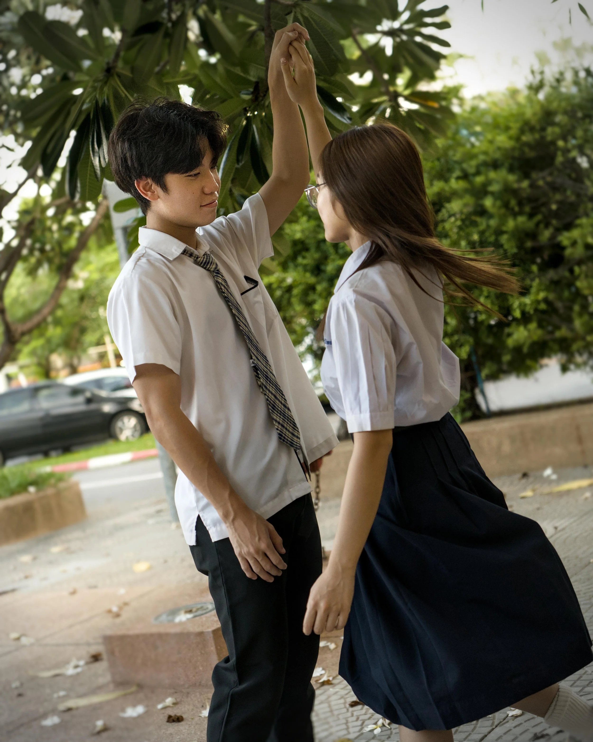 A young man and young woman are standing close together outdoors, holding hands up above their heads, with the young man looking at the young woman. They are wearing school uniforms, and the background shows trees and parked cars.