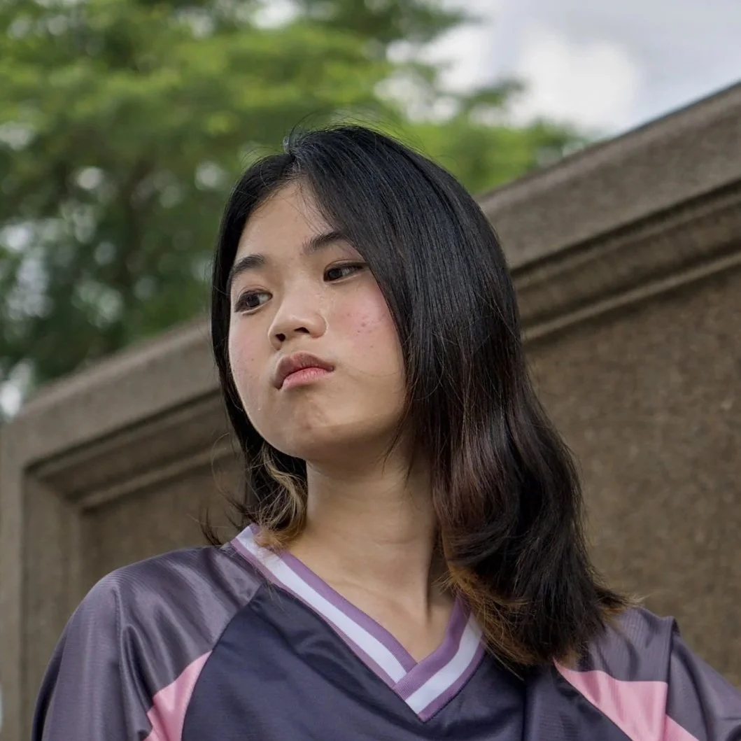 A young woman with black hair, wearing a purple and pink sports jersey, standing outdoors next to a stone or concrete wall with green trees in the background.