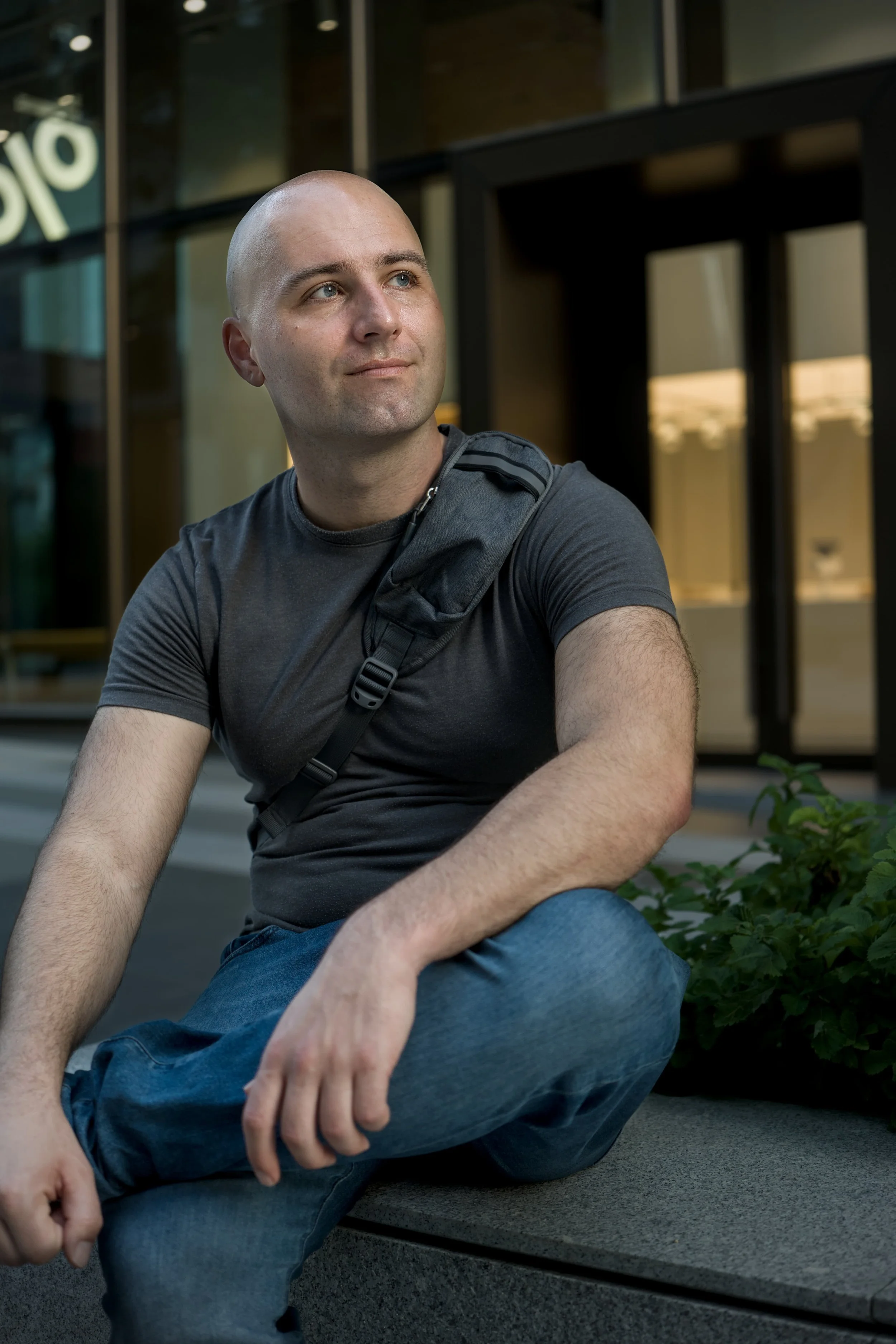 A young man with a shaved head and light skin sitting outdoors on a black bench next to a building with glass windows. He is wearing a dark gray t-shirt and blue jeans, with a black strap over his shoulder. The background shows some greenery and a pa