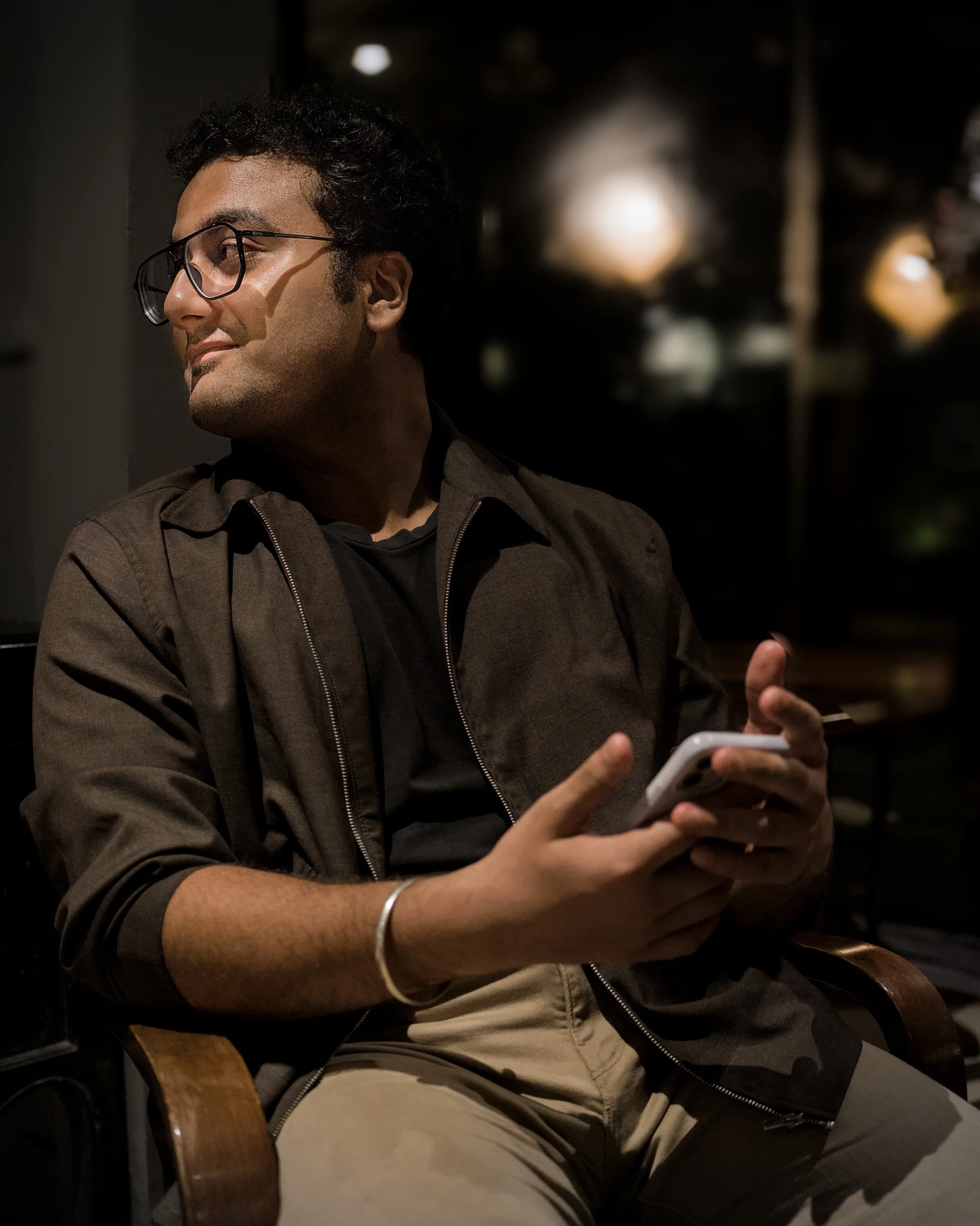 A young man with glasses, dark hair, and a clean-shaven face is sitting in a dimly lit indoor space, looking to his left while holding a smartphone in his hands. He is wearing a dark jacket over a black shirt, and beige pants. The background is blurr