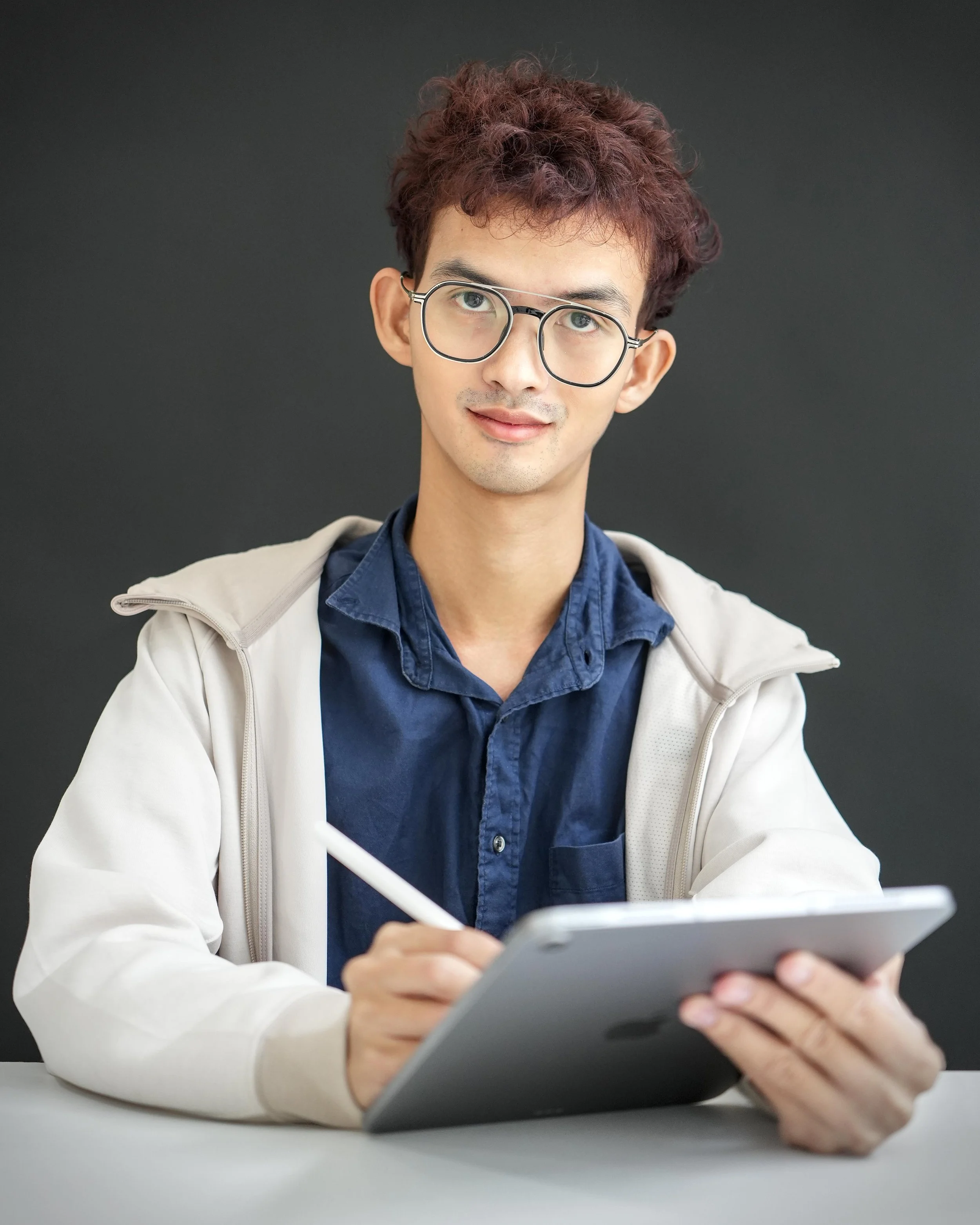 A young man with curly hair, glasses, and light skin wearing a blue shirt and light jacket, holding a tablet device, sitting at a desk against a dark background.