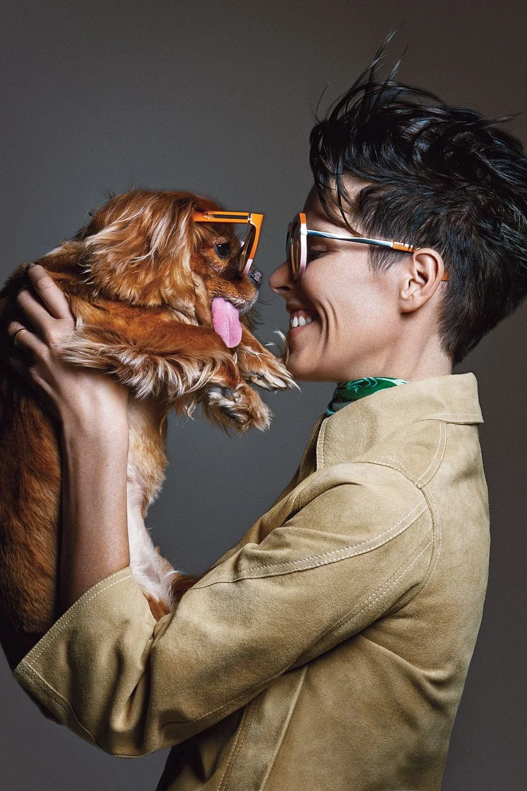 A woman with short black hair and glasses holding a small brown dog with glasses, nose, and tongue out, facing each other close, smiling, in a studio with a plain gray background.