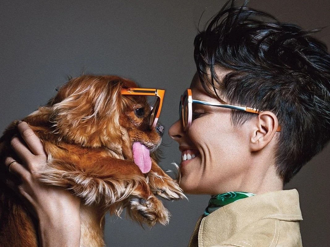 A woman holding a small brown dog wearing glasses face-to-face, both with noses touching, in a close-up portrait.