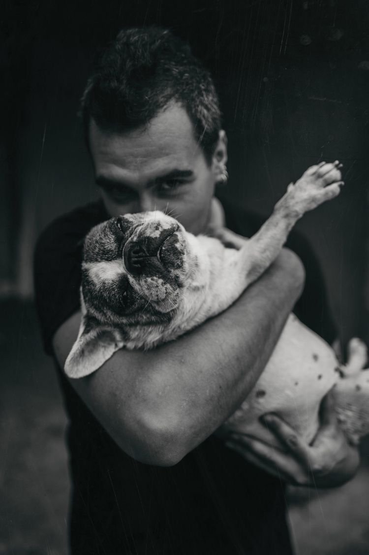 A person with short dark hair holding a small dog, possibly a bulldog, in their arms. The dog appears to be relaxed with its eyes closed and is being embraced in a gentle hug. The image is in black and white.