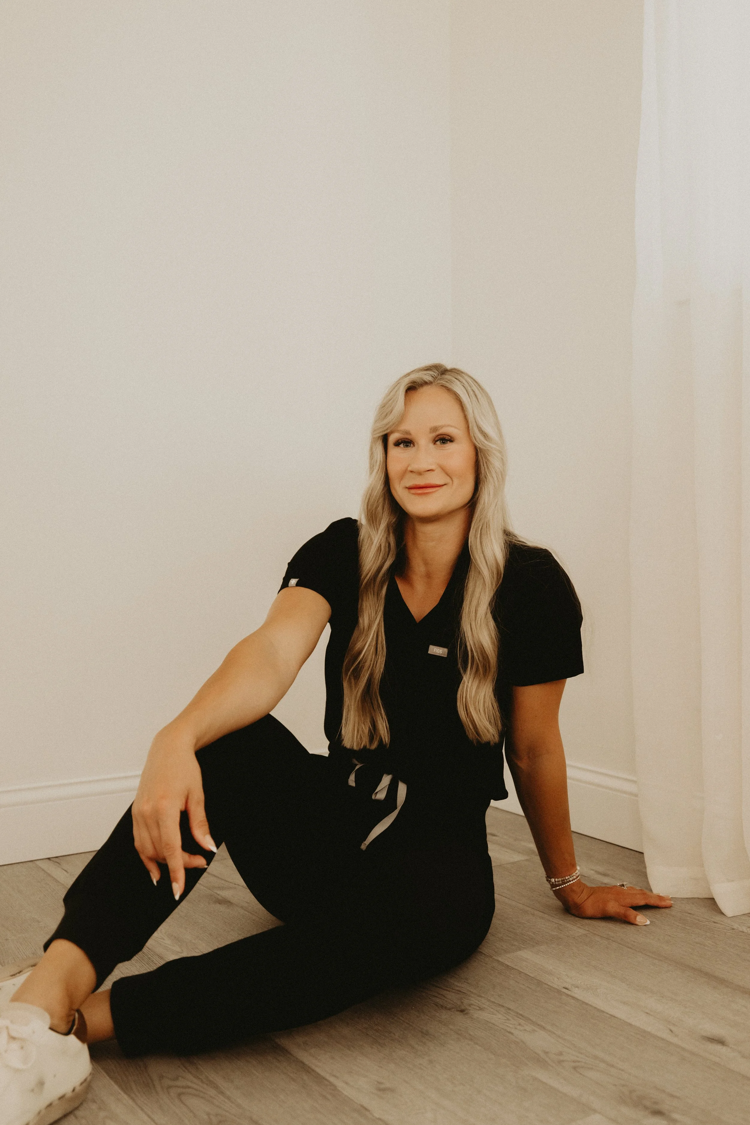 A woman with long blonde hair sitting on the wooden floor in a black outfit, smiling, with one arm resting on her bent knee and the other hand on the floor, against a plain white wall.