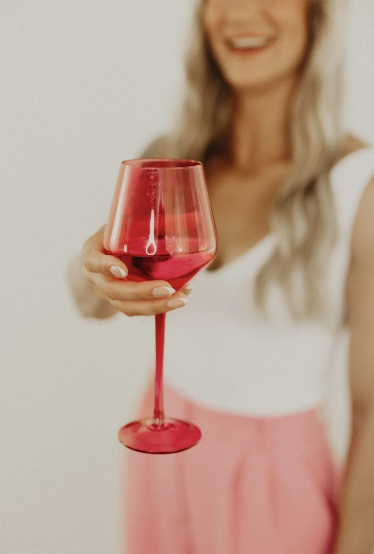 A woman with blonde hair and a white top holding a glass of red wine, smiling, with a blurred background.