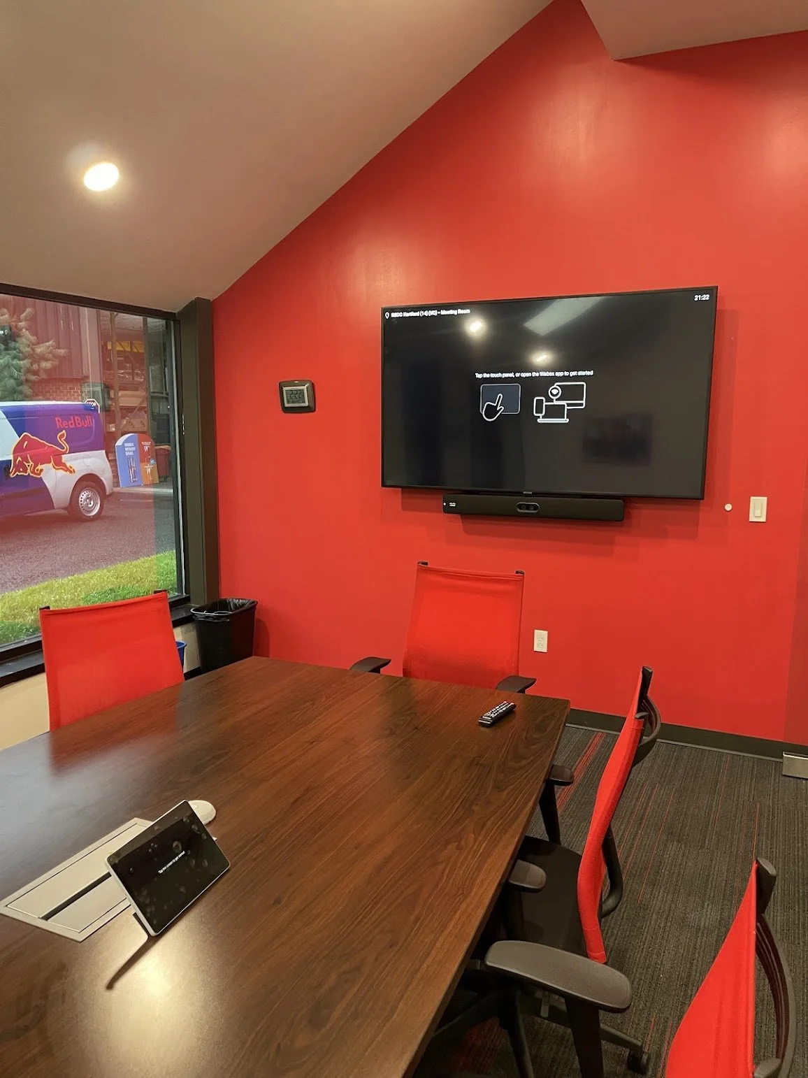 A conference room with a large wooden table surrounded by red chairs, a wall-mounted TV screen with instructions, a remote control, a tablet on the table, and a window showing a Red Bull truck outside.