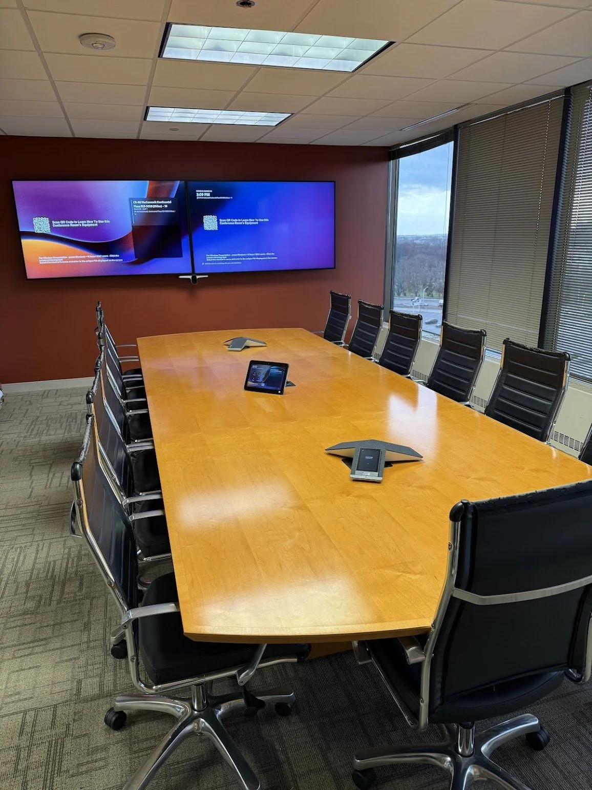 Empty conference room with a large wooden table, black office chairs, wall-mounted display screens showing a QR code, and windows with blinds overlooking a cityscape.