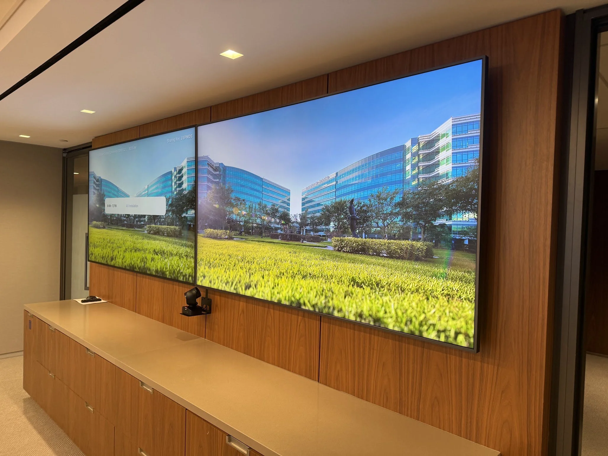 Large digital display screen showing an image of modern office buildings with glass exteriors, green trees, and a blue sky, mounted on a wooden wall in an indoor setting.