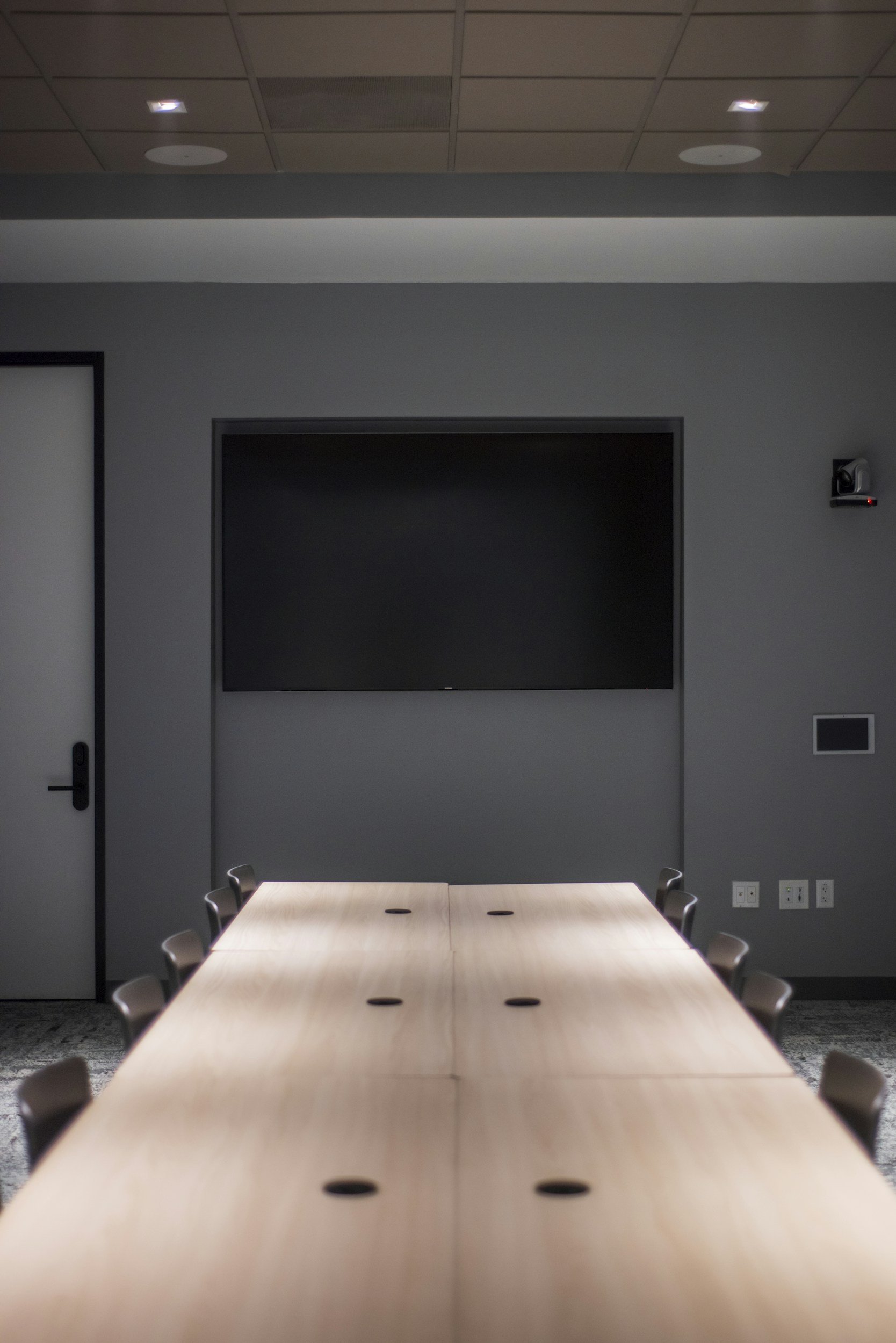 A conference room with a long wooden table and chairs, a large black screen on the wall, and a ceiling with lights.