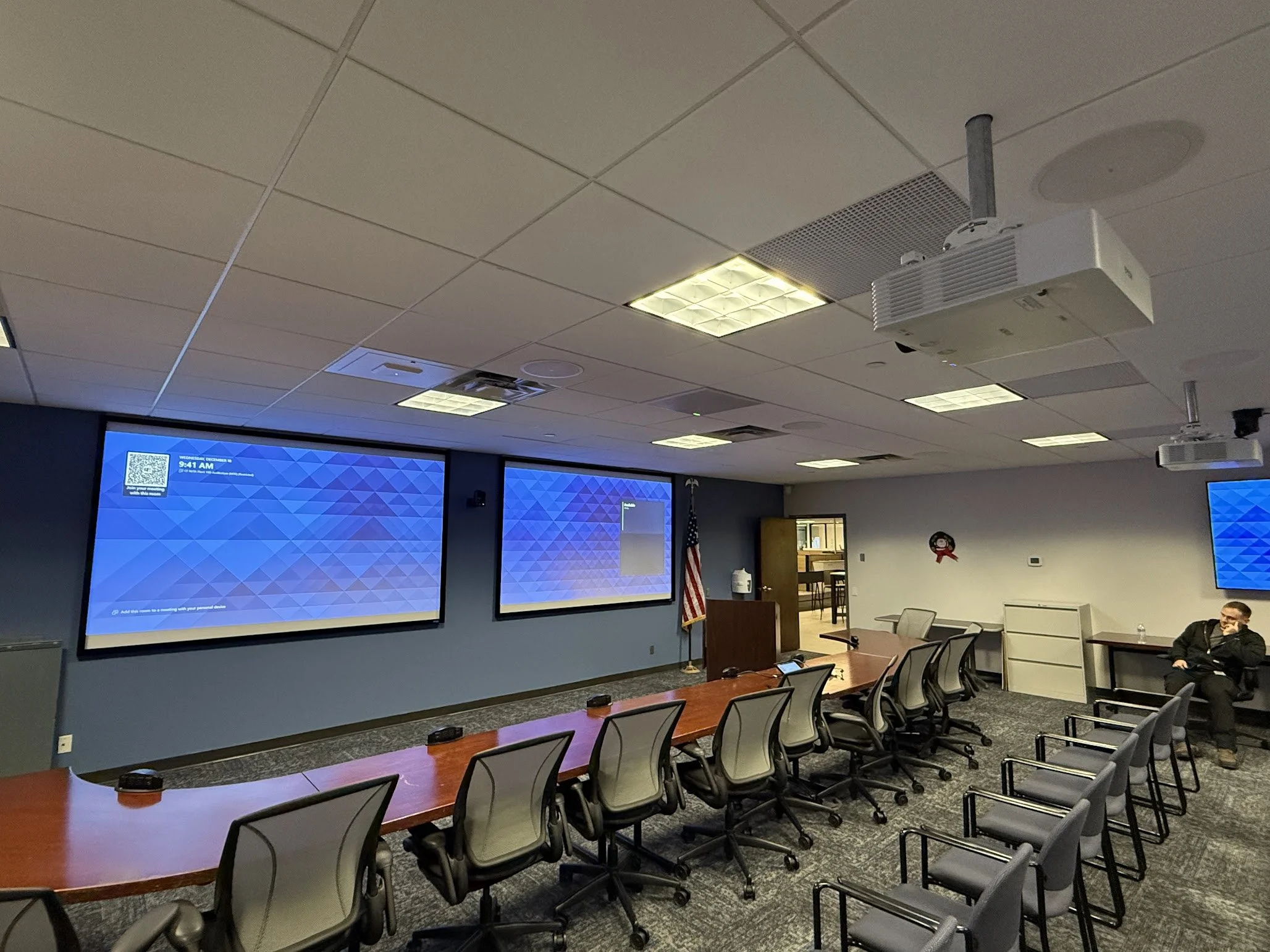 A conference room with a long wooden table, office chairs, and three large screens on the wall displaying a blue geometric background. There are people sitting at the table and in the background, with a flag and some office furniture.