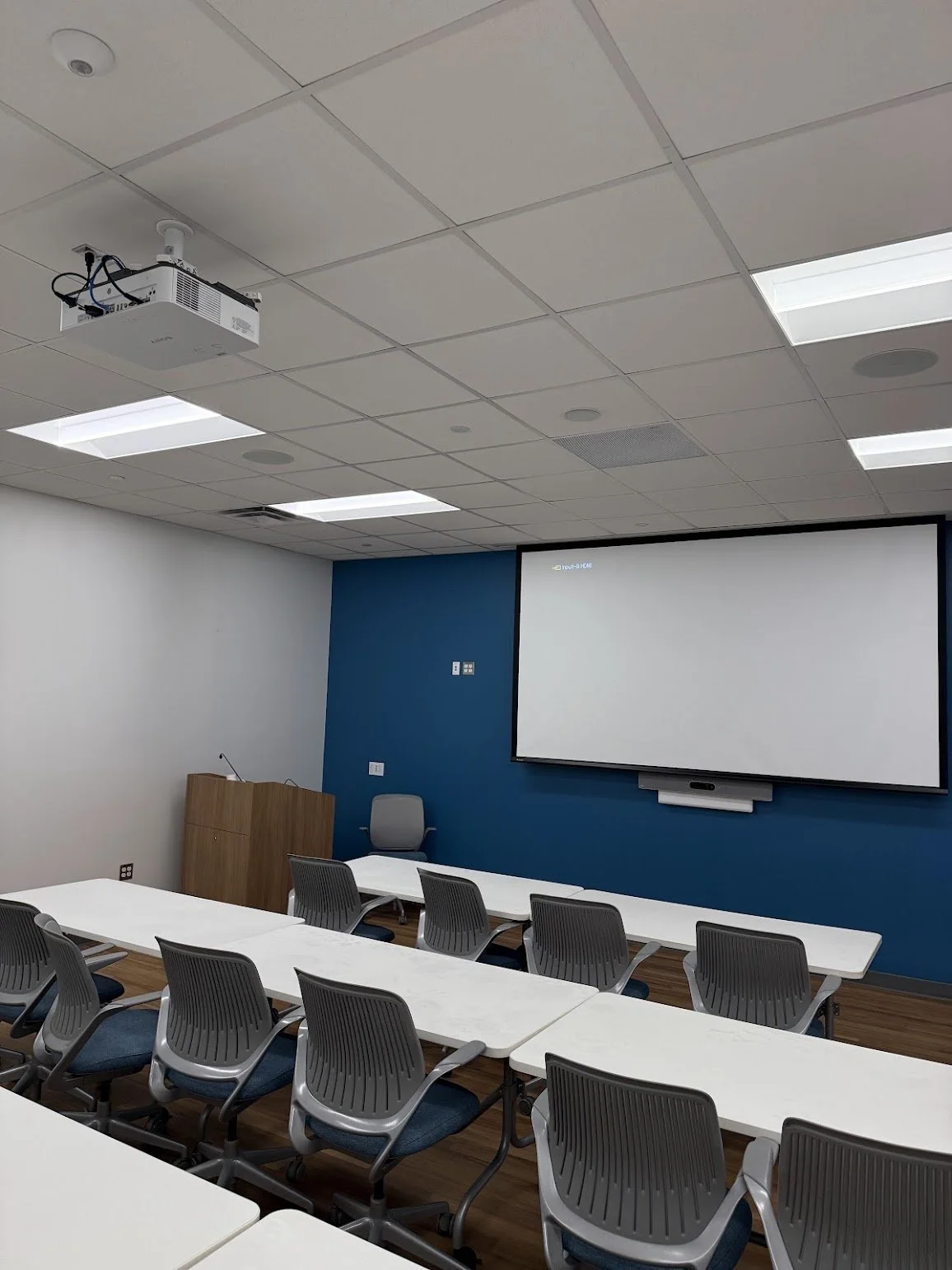 An empty classroom or conference room with white desks, gray chairs, a projector mounted on the ceiling, and a large screen at the front, with a blue accent wall and a wooden podium.