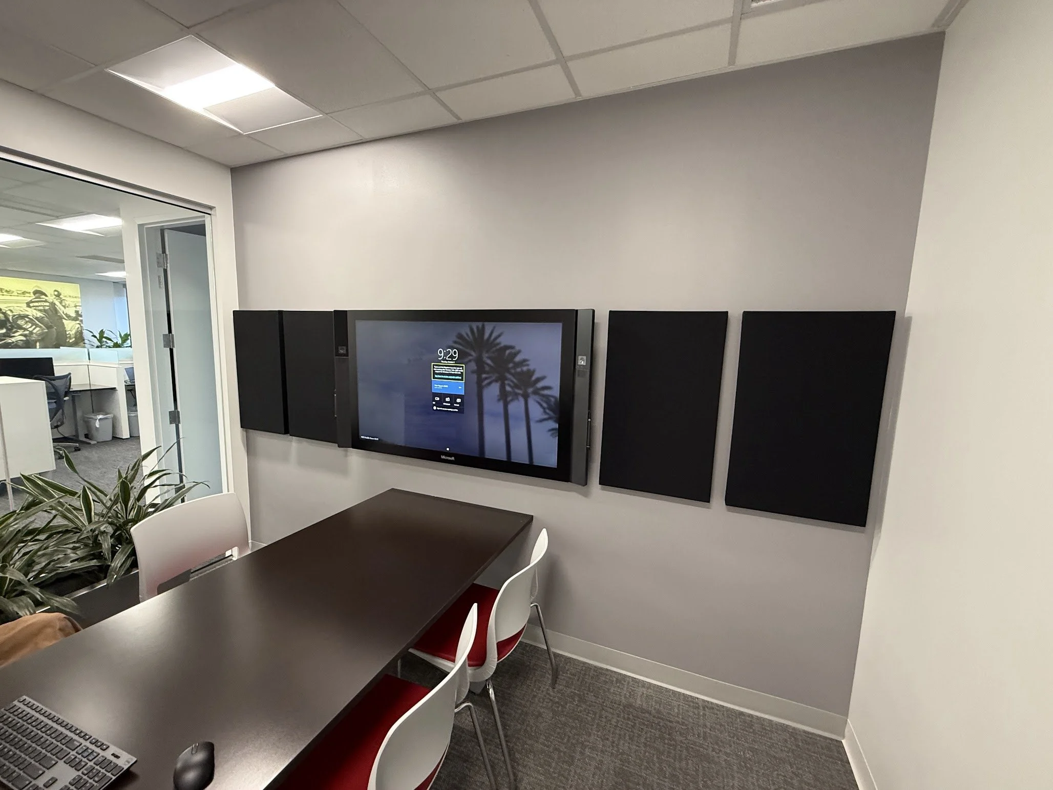 An office room with a television screen mounted on the wall displaying a scene of palm trees at sunset or sunrise. There is a black table and white chairs nearby, a woman sitting at the table, and some office furniture visible in the background throu