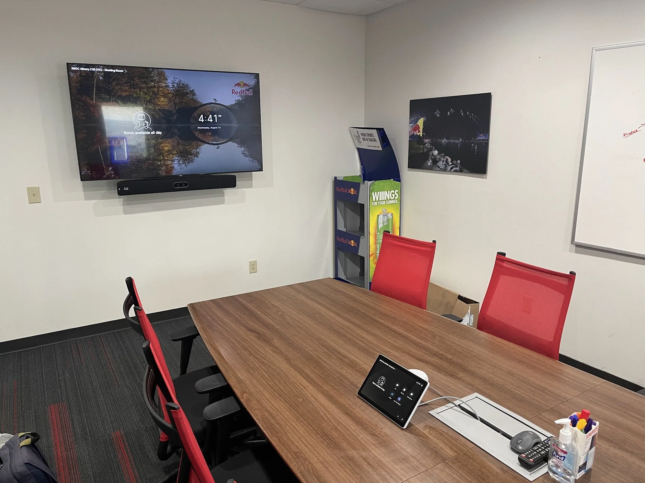 A meeting room with a wooden table surrounded by red chairs, a wall-mounted large screen displaying a nature scene with trees and a lake, and a whiteboard. Items on the table include a tablet, a keyboard, a mouse, a container with markers, hand sanit