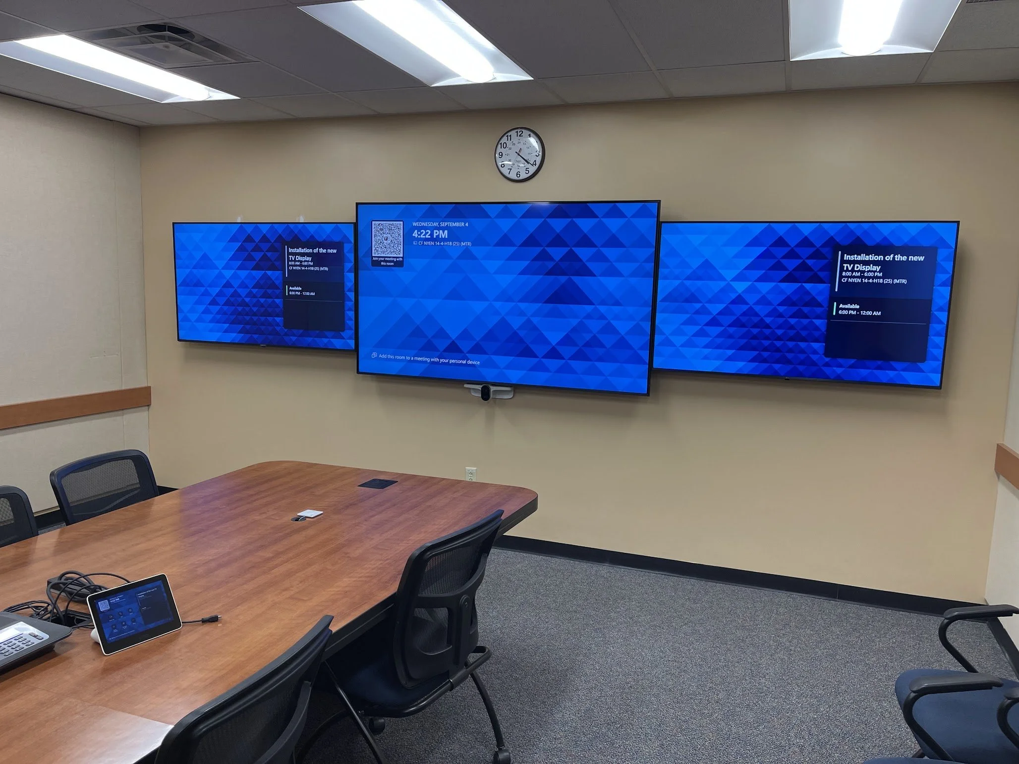 A conference room with a large wooden table and black chairs. On the table, there is a phone, a tablet, and some cables. On the beige wall, there are three flat-screen monitors and a round clock showing 4:20. The ceiling has fluorescent lights.