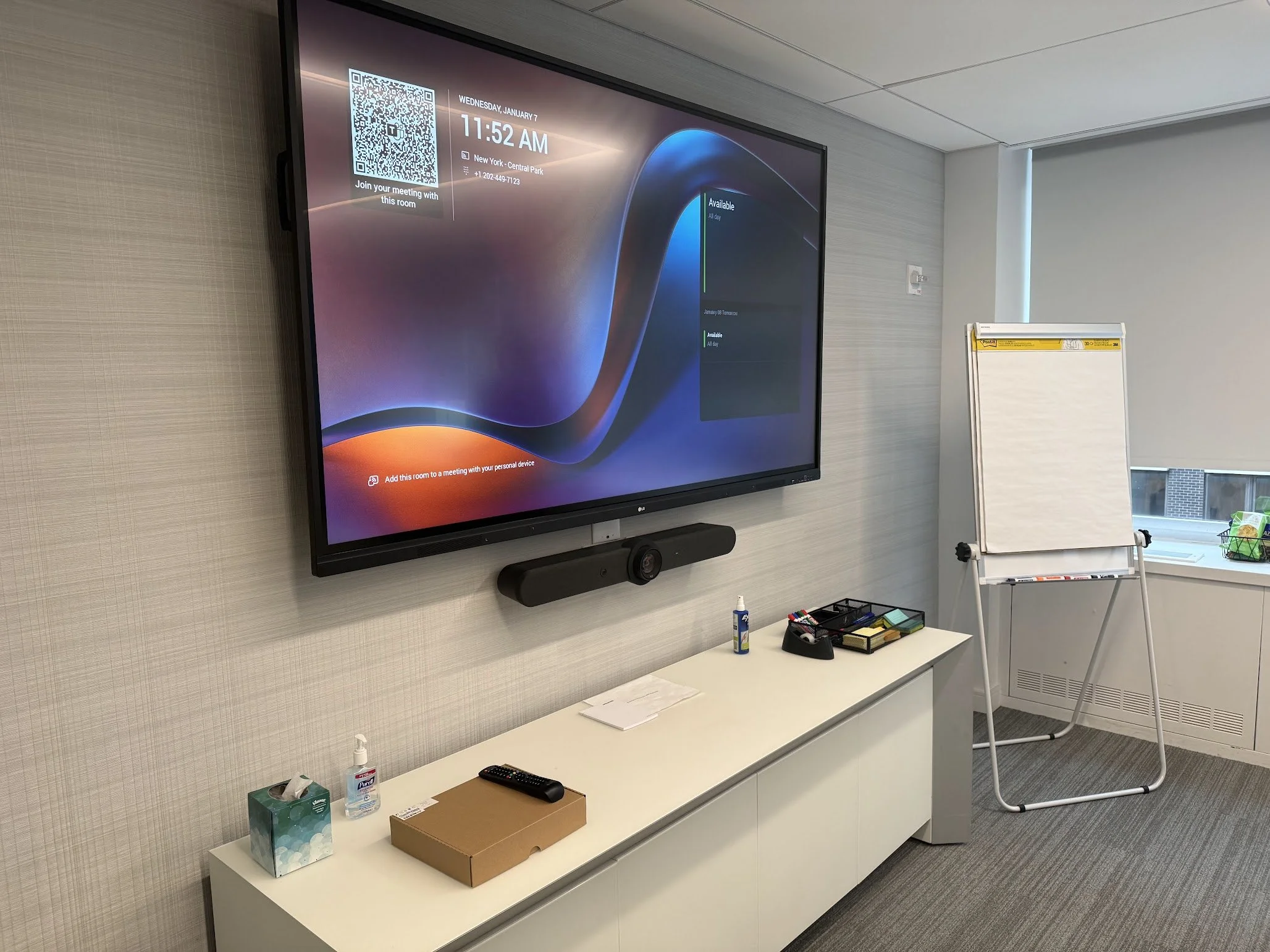 A digital conference room display showing the date, time, location, and a QR code for joining a meeting. The room setup includes a whiteboard on a stand, various items on a white counter, including a tissue box, hand sanitizer, a remote, and some box