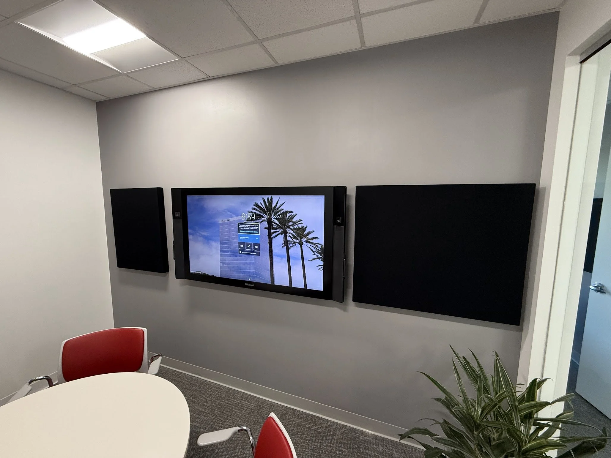Office conference room with a flat screen monitor, surrounding chairs, a potted plant, and part of a door visible.