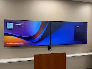 A large flat-screen monitor displaying a split screen with two sections, mounted on a wall in a conference room, with a wooden podium below it.