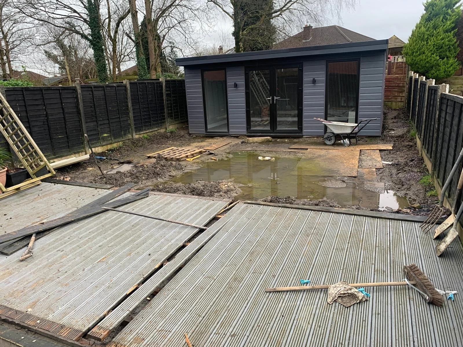 Backyard undergoing renovation with partially installed decking, a muddy area with standing water, a small grey shed, and construction tools and materials.